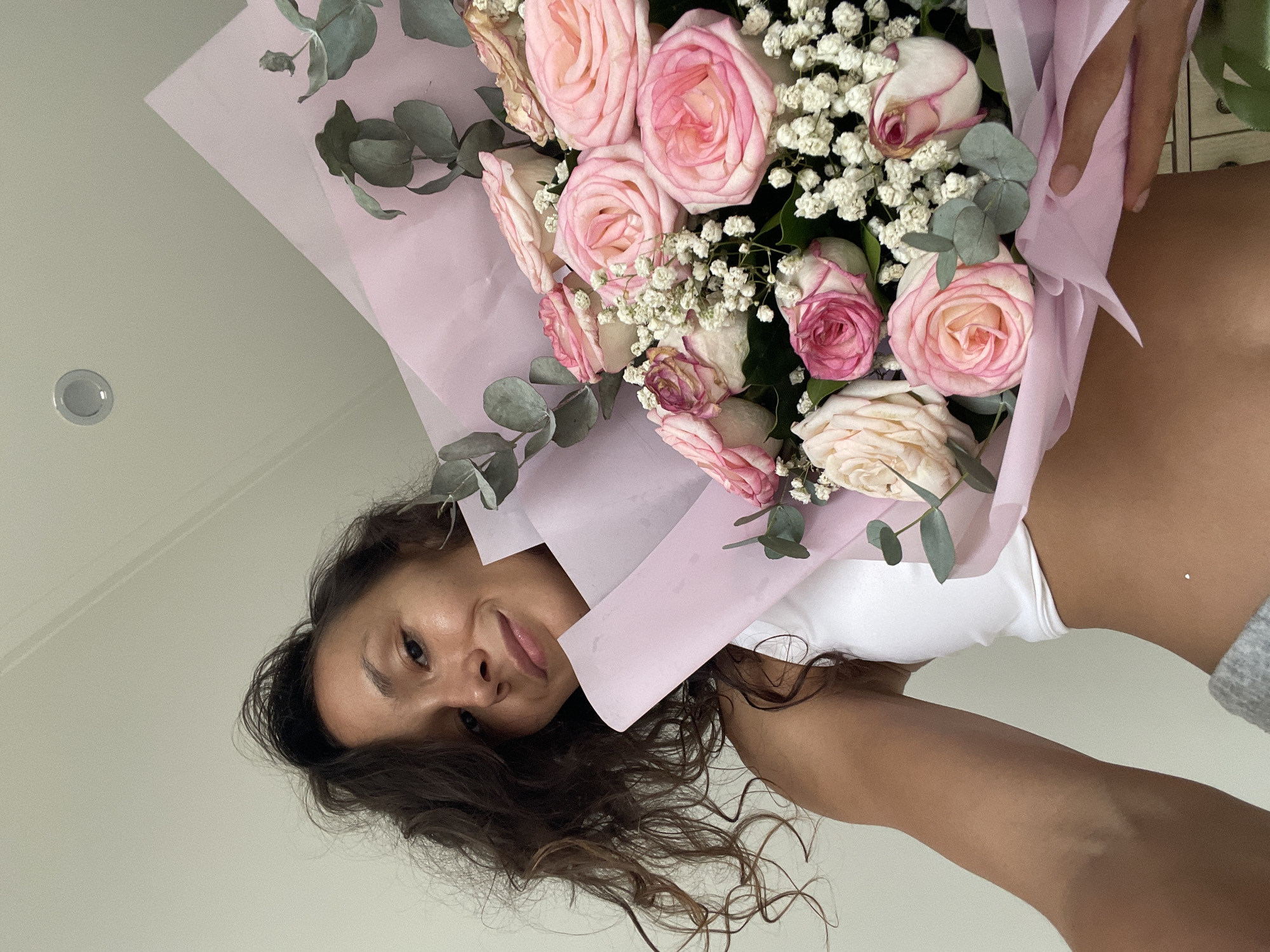 In Phu Thuy Ward, Vietnam, a young woman with curly dark brown hair is holding a bouquet of pink and white roses. She is wearing a white sports bra and grey sweatpants. The bouquet is wrapped in pink paper and accented with eucalyptus leaves and baby's breath. The woman is looking at the camera with a slight smile. The background appears to be an indoor setting with a white wall and ceiling. The image is taken from a low angle, looking up at the woman.