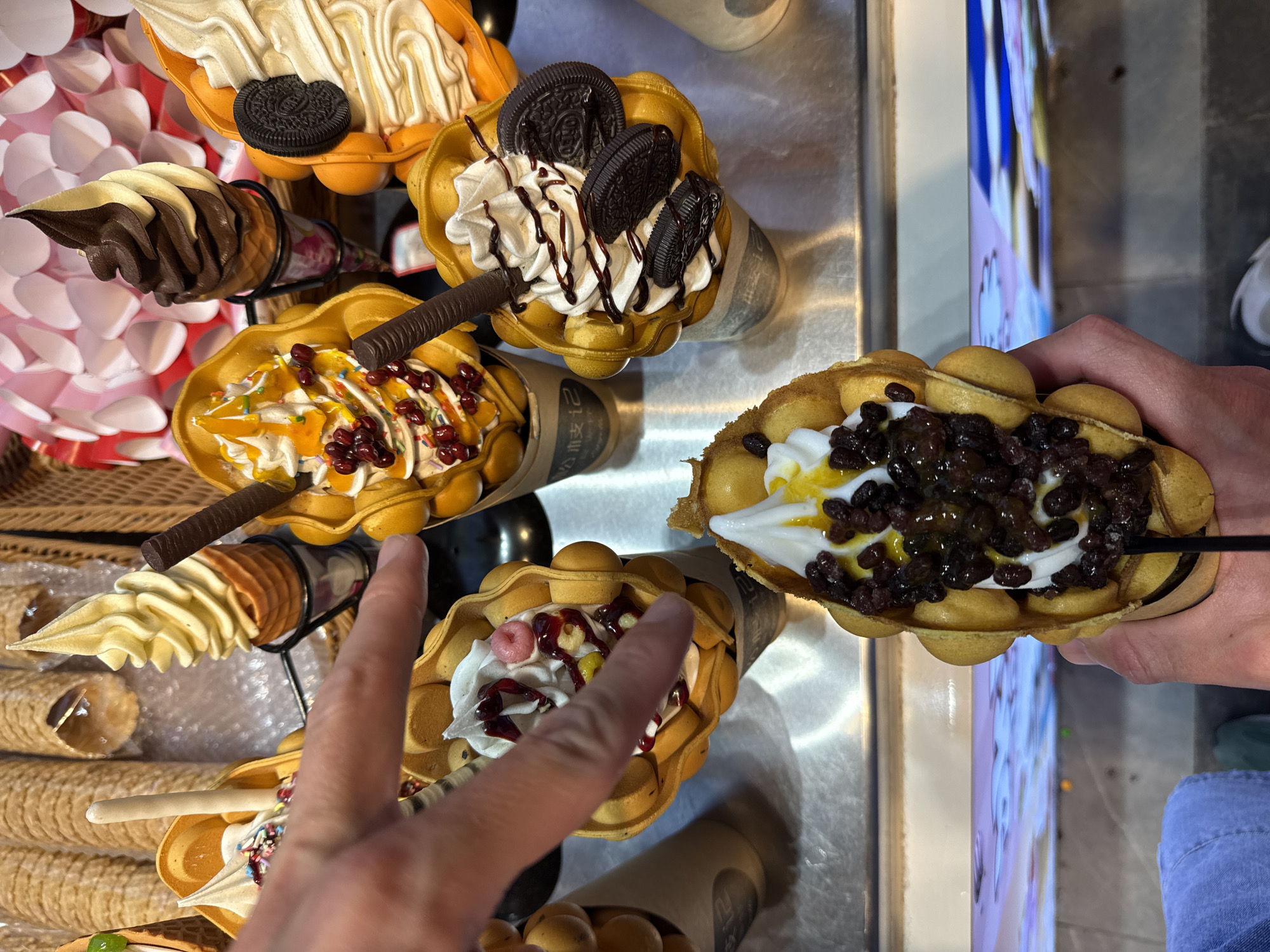 A slightly high-angle, medium shot depicts a collection of egg waffle desserts, also known as gai daan zai, displayed on a metallic surface. A right hand, out of focus, extends from the left into the frame, gesturing towards the desserts. A second hand appears on the right side of the image holding a dessert. The desserts vary in toppings and fillings, including ice cream, chocolate, fruit, sprinkles, and what appear to be Oreo cookies. The desserts are served in cone-shaped paper containers with visible Asian script. The setting suggests a dessert stall or shop, possibly located in the Furong district of Changsha, China, as gai daan zai are commonly found there.