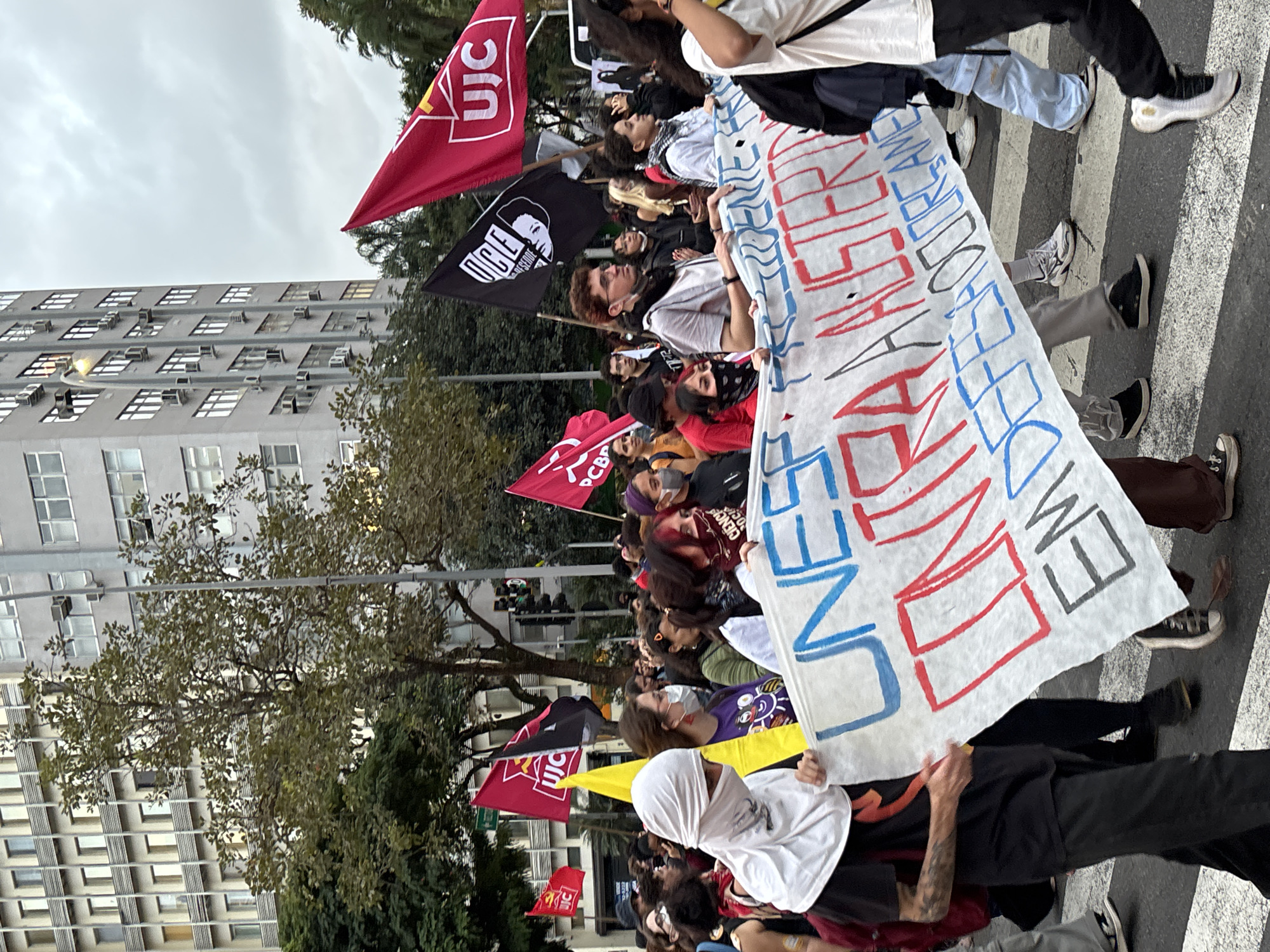 A street-level shot shows a protest march in São Paulo, Brazil. A multi-story building is visible in the background. The weather is overcast. Protesters hold up a banner with the acronym "UNE" written on it in red and blue letters. Various red flags with the letters "UIC" are visible among the protesters, as well as a black flag. Protesters are standing on a pedestrian crosswalk. The photo is taken from street level, showing the crowd moving along the street.