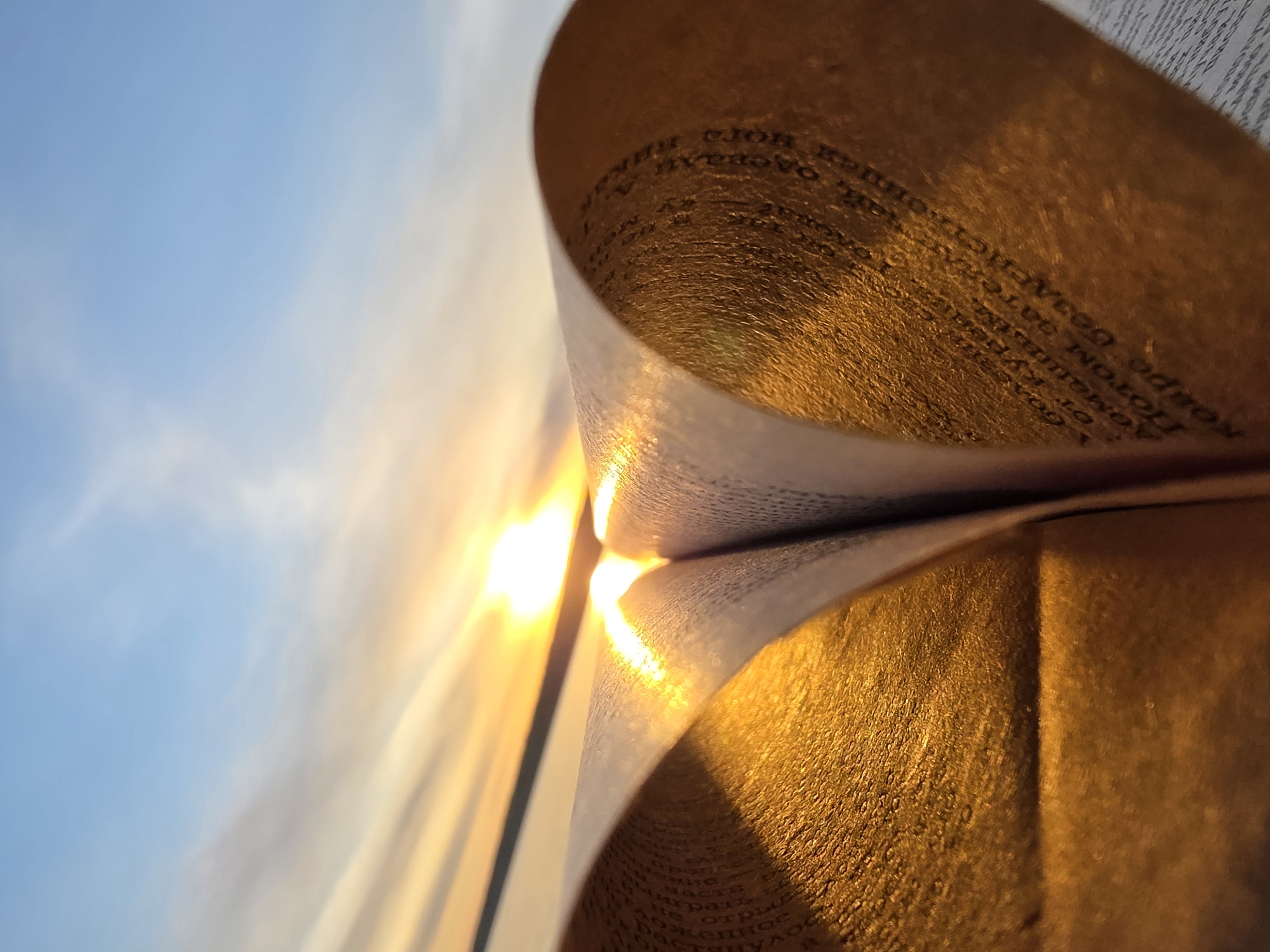 A close-up perspective of a book held open against a sky containing sunlight and light clouds. The book's pages are curved and the light shines through them, creating a radiant glow. The sky is blue and partially obscured by the book. The text printed on the book's pages is visible. The scene likely depicts an outdoor setting given the presence of natural light and the sky. The location is Chaikovsky, Russia.
