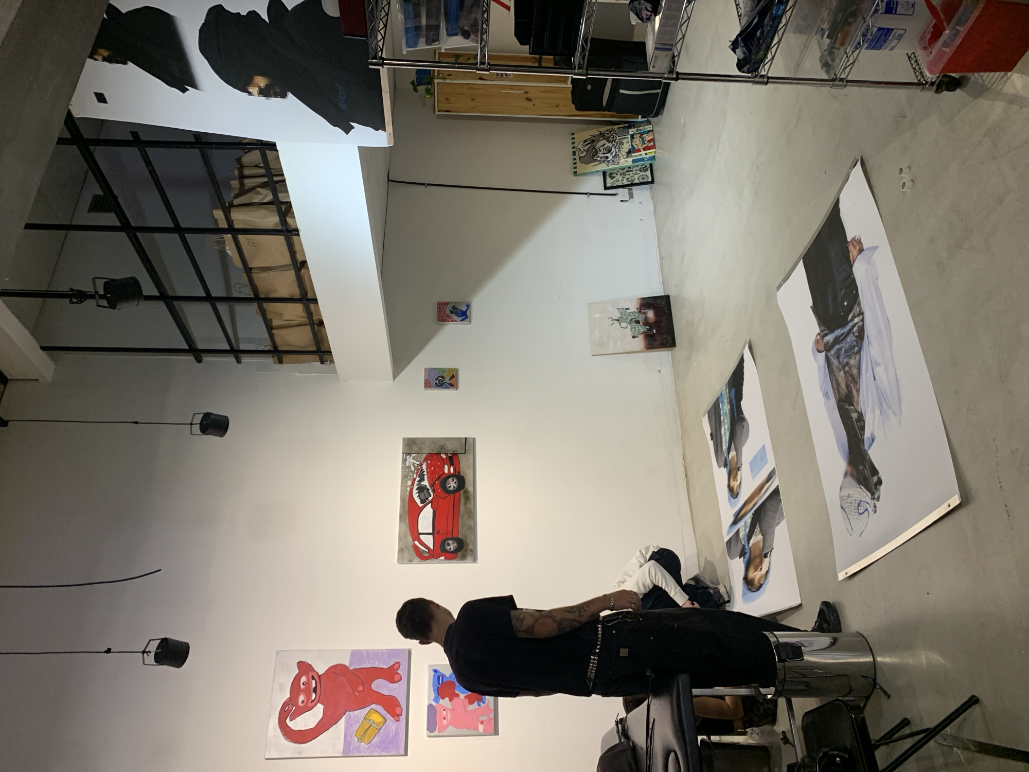 The image depicts an indoor space, likely an art gallery or studio, with exposed ceilings and an industrial aesthetic. The location is suggested to be Buenos Aires, Argentina, although there are no explicit indicators of location within the image. The walls are primarily white and feature an assortment of framed artwork, including abstract pieces, a red car, and a red figure. Larger, unframed canvases lean against the wall on the floor, appearing to be portraits. A man is seen in the foreground, dressed in black, standing near what seems to be a silver metal container, possibly a trash can. The setting is illuminated by a combination of natural light and track lighting fixtures. There are racks with clothing and miscellaneous objects in the background. A chair and other loose objects are scattered throughout the space.