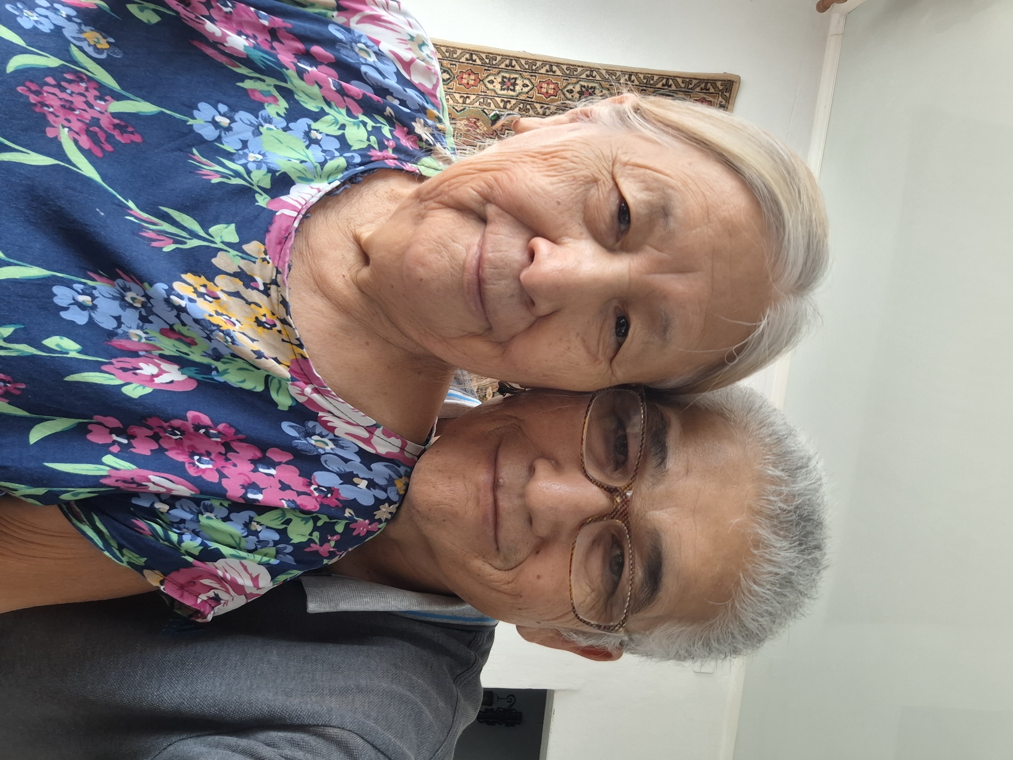 Two elderly individuals, a woman and a man, are captured in a close-up selfie, likely in a home environment in Almaty, Kazakhstan, given the background tapestry which is characteristic of Central Asian decor. The woman, on the left, has fair skin with wrinkles, graying blonde hair, and is wearing a colorful floral blouse with a dark blue background. She is looking directly at the camera with a gentle smile. The man, on the right, has fair skin, gray hair, and is wearing glasses with a patterned frame. He is wearing a gray collared shirt and is also smiling and looking towards the camera. They appear to be a couple, with the woman leaning slightly towards the man. The background is a softly lit room with a white wall and a tapestry with geometric patterns in earthy tones hanging on the left. A doorway is visible in the background on the right.