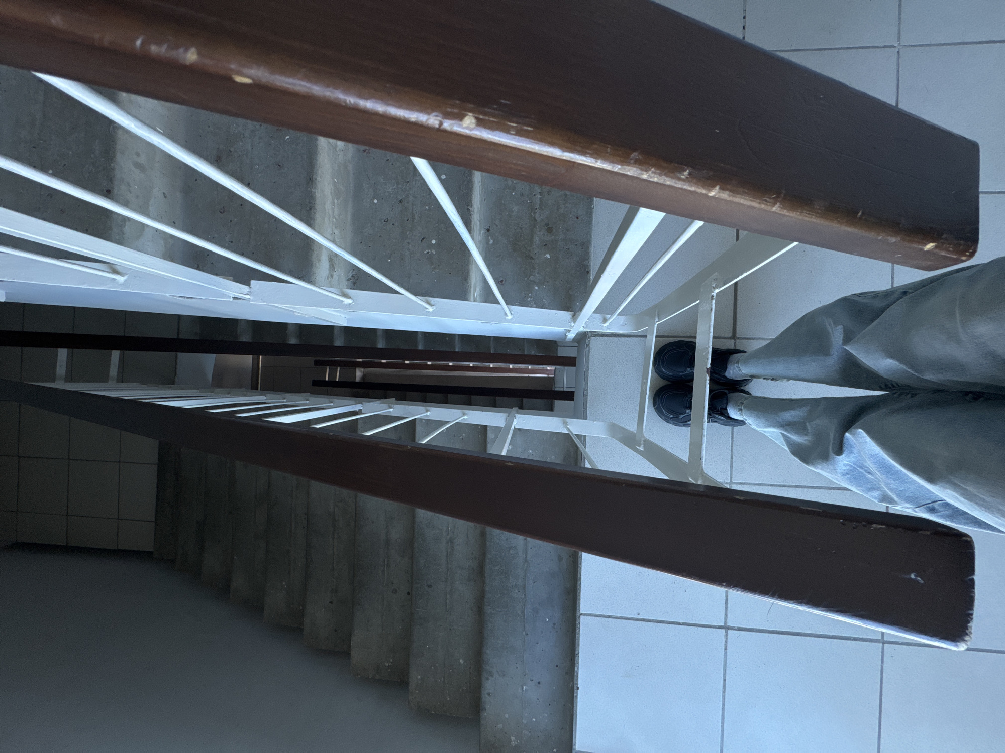 An overhead view captured from a multi-story stairwell in Chaikovsky, Russia, shows a person's legs and feet positioned on a light grey tiled landing. The individual wears light blue jeans and dark athletic shoes. The perspective looks directly down into the stairwell, revealing multiple flights of grey concrete stairs descending. Two dark brown wooden handrails frame the central void, with white metal balusters extending from the right-side handrail. The concrete steps exhibit visible wear and scattered dark specks. The walls of the stairwell are a plain grey, and the lower sections of the descent appear less illuminated than the immediate landing. No other individuals or direct interactions are visible.
