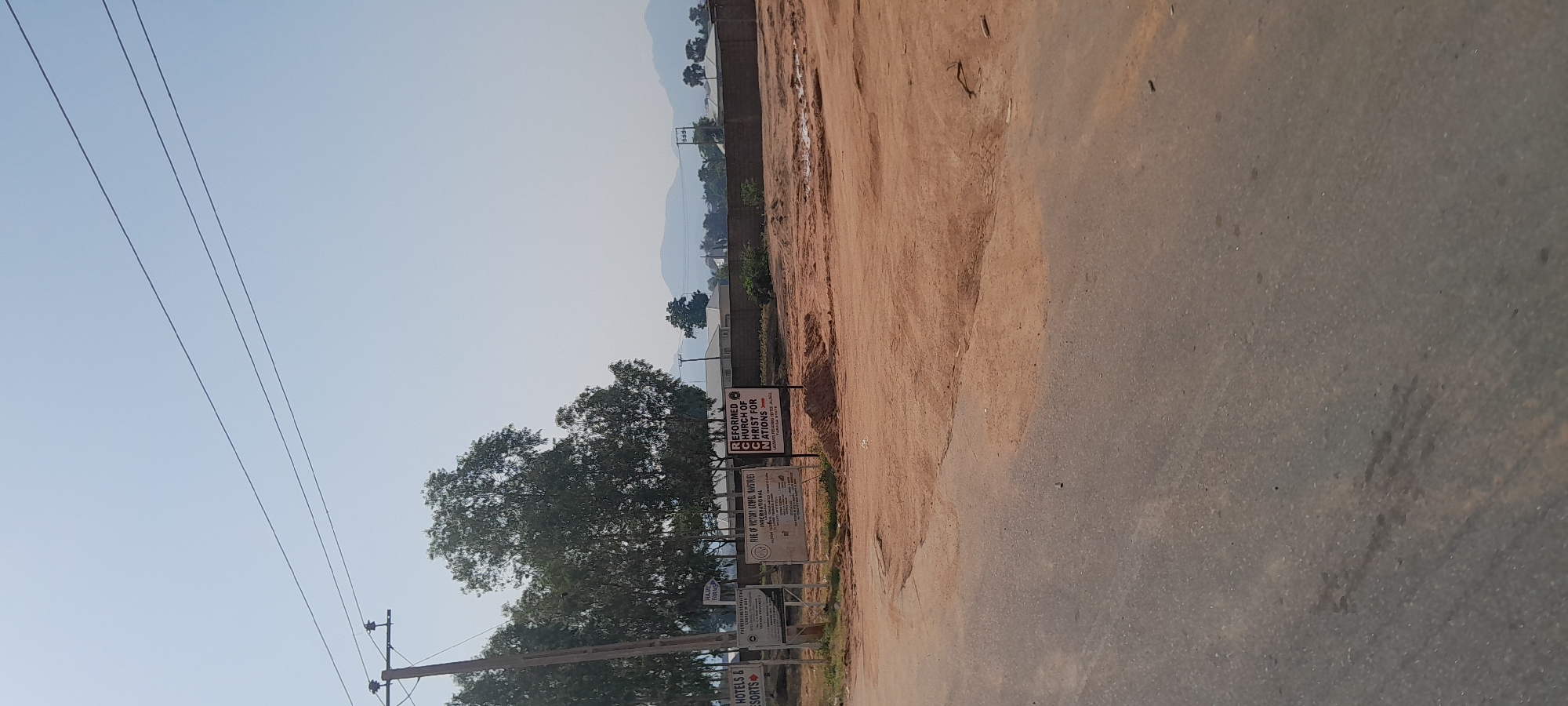 The image shows a dirt road with some signs and trees on the side. The road appears to be in an undeveloped area. The sky is clear and blue. In the background, there are some buildings and mountains. The scene is likely in Jalingo, Nigeria, as indicated by the signage. One sign says "REFORMED CHURCH OF CHRIST FOR NATIONS" and below that "JALAMBE PRESBYTERIAN CENTER, JALINGO, TARABA STATE". Another sign reads "FIRE OF VICTORY GOSPEL MINISTRIES INTERNATIONAL". A third sign is partially visible and appears to be for hotels and resorts.