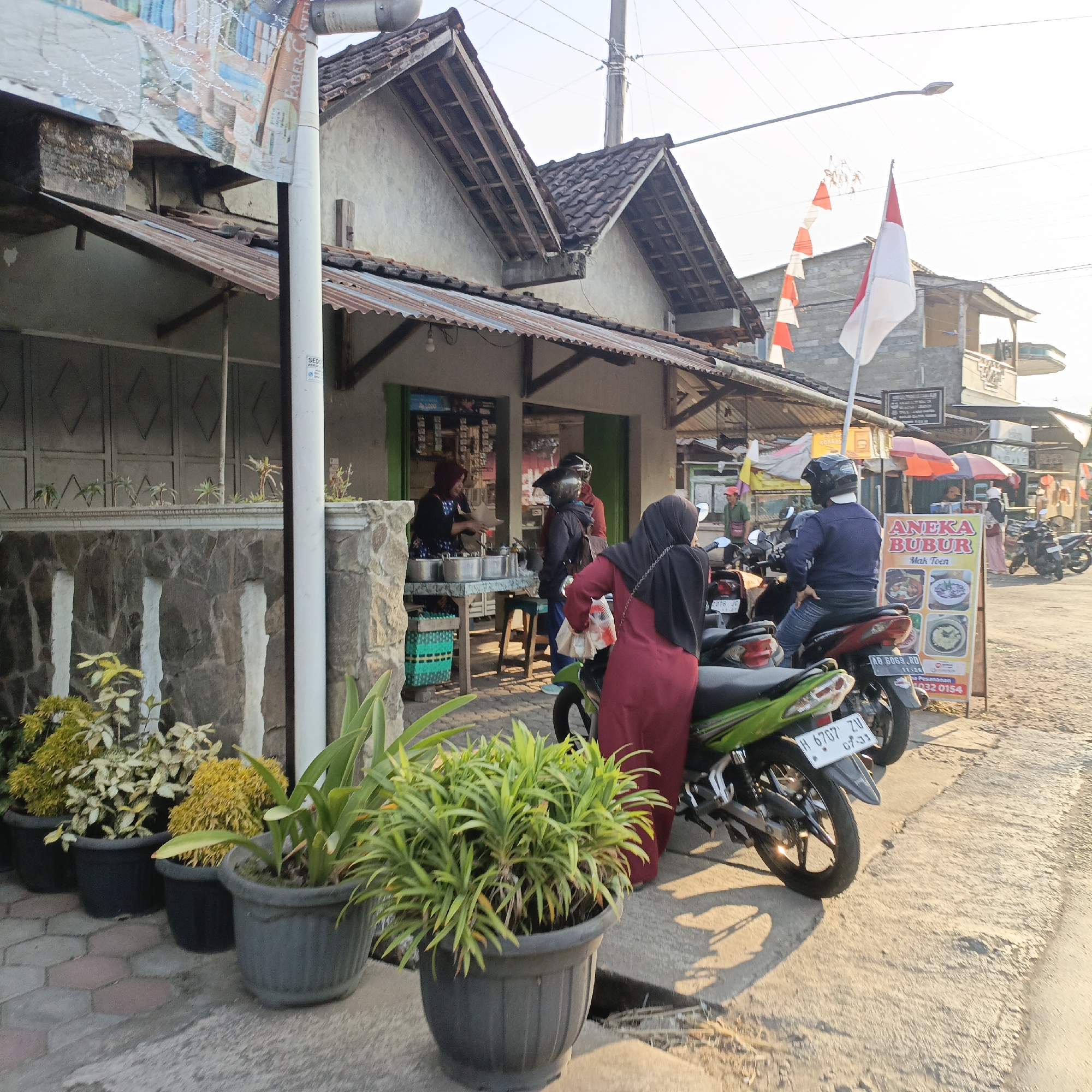 This is a medium shot, taken outdoors in Sraten, Indonesia, during the daytime with the sun casting long shadows. The scene depicts a street-side food stall with several people and motorcycles.
In the foreground, to the left, are several potted plants. The plants in the closest pots are leafy green, while to their left are plants with yellowish-green foliage.
The main focus is a food stall, possibly a bubur (porridge) stall, indicated by a sign that reads "ANEKA BUBUR" and shows pictures of food. The stall has a covered awning supported by wooden beams. A table in front of the stall holds various metal containers, likely for food ingredients.
Several people are present. A woman in a black headscarf and a maroon-colored dress is standing at the stall, appearing to purchase or receive food. Another person, also wearing a headscarf and maroon clothing, is partially visible leaning against a green motorcycle. A person wearing a blue shirt, jeans, and a helmet is seated on a red motorcycle to the right of the stall. To the left of the stall, a woman in a dark headscarf and patterned apron is preparing food, with several metal pots in front of her. Behind the stall, a man in a green shirt and a red cap is walking away from the scene.
Motorcycles are parked along the roadside. The green motorcycle in the foreground has a license plate that reads "H 6767 ZN". A red motorcycle is parked next to it.
The environment includes a paved road, a concrete sidewalk, and the buildings of the food stall. A flagpole with a red and white Indonesian flag is visible behind the stall, along with a decorative string of red and white pennants. A street lamp is positioned above the scene. The background shows more buildings, some with tiled roofs, and power lines. The overall atmosphere suggests a typical street scene in a residential or commercial area.