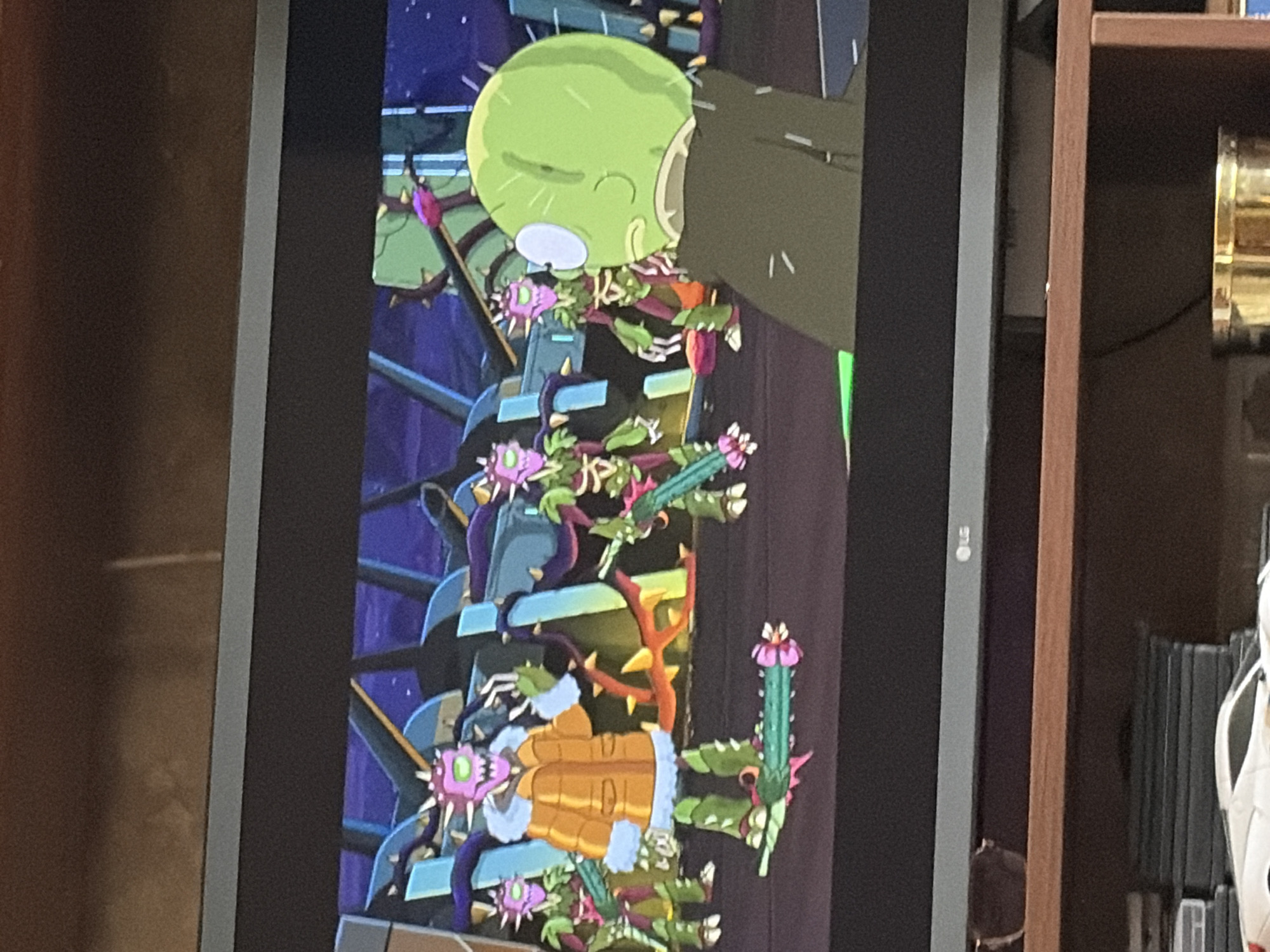 The image displays a scene from an animated television show, likely Rick and Morty, projected on a television screen. In the foreground, a character with a green, bulbous head and a dark cloak is seen in profile, facing away from the viewer. Behind this character, a group of alien creatures with purple, spiky heads, green skeletal bodies, and four arms are visible. One of these aliens is prominently positioned in the center, wearing an orange coat with a fur-lined collar and hood. It holds a spiky, green weapon in its hands. The background appears to be an indoor setting with futuristic architecture, characterized by blue and purple structures and lighting. The scene is set in an unknown urban environment.
