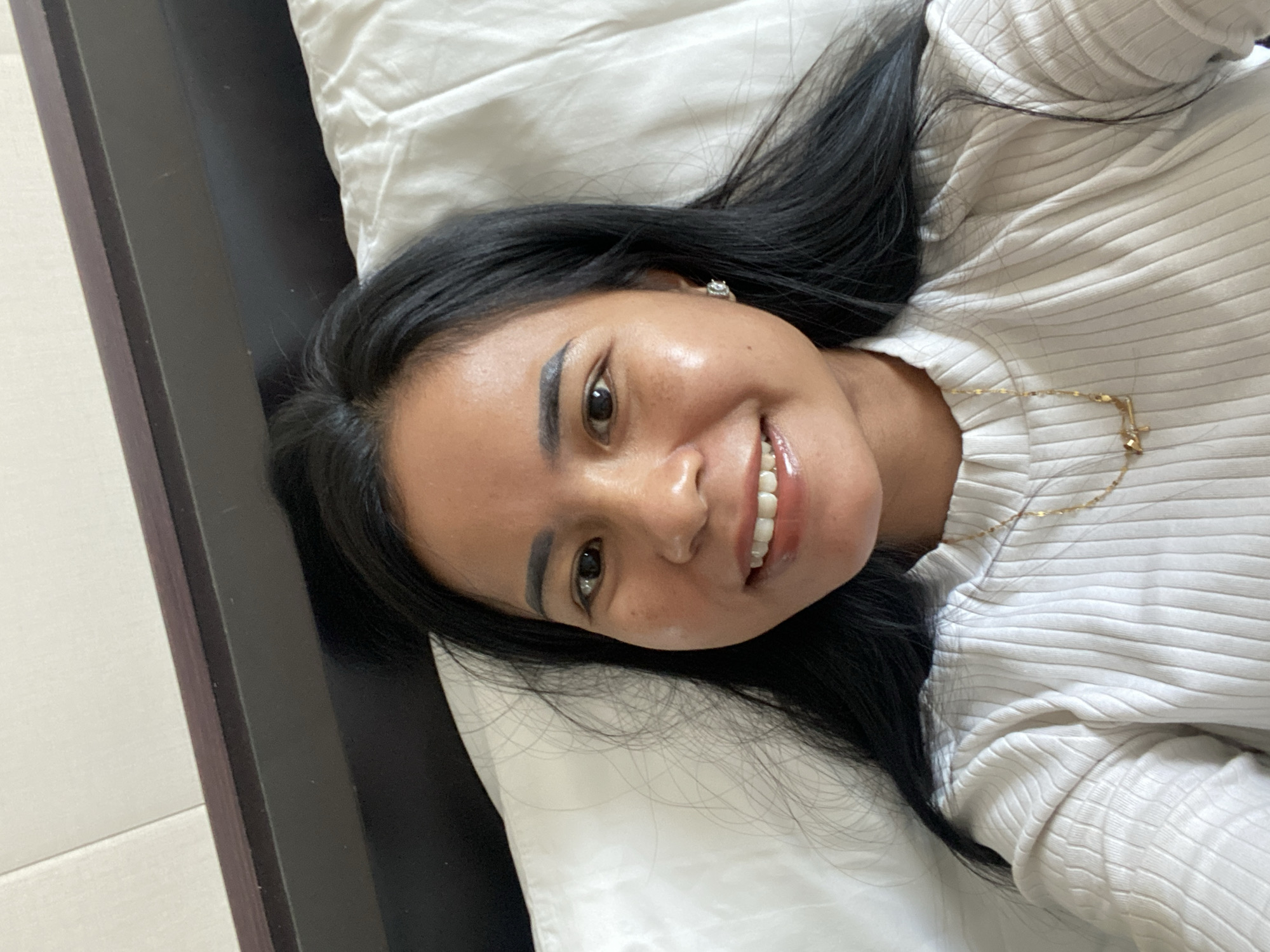 A woman with dark hair and fair skin lies on a white pillow in a horizontal pose. She is wearing a light-colored, ribbed, turtleneck sweater and a delicate gold necklace. She is smiling, and her gaze is directed towards the viewer. The background includes a dark-colored bed frame and a light-colored wall. The setting appears to be indoors, and the woman's location is North Kuta, Indonesia.