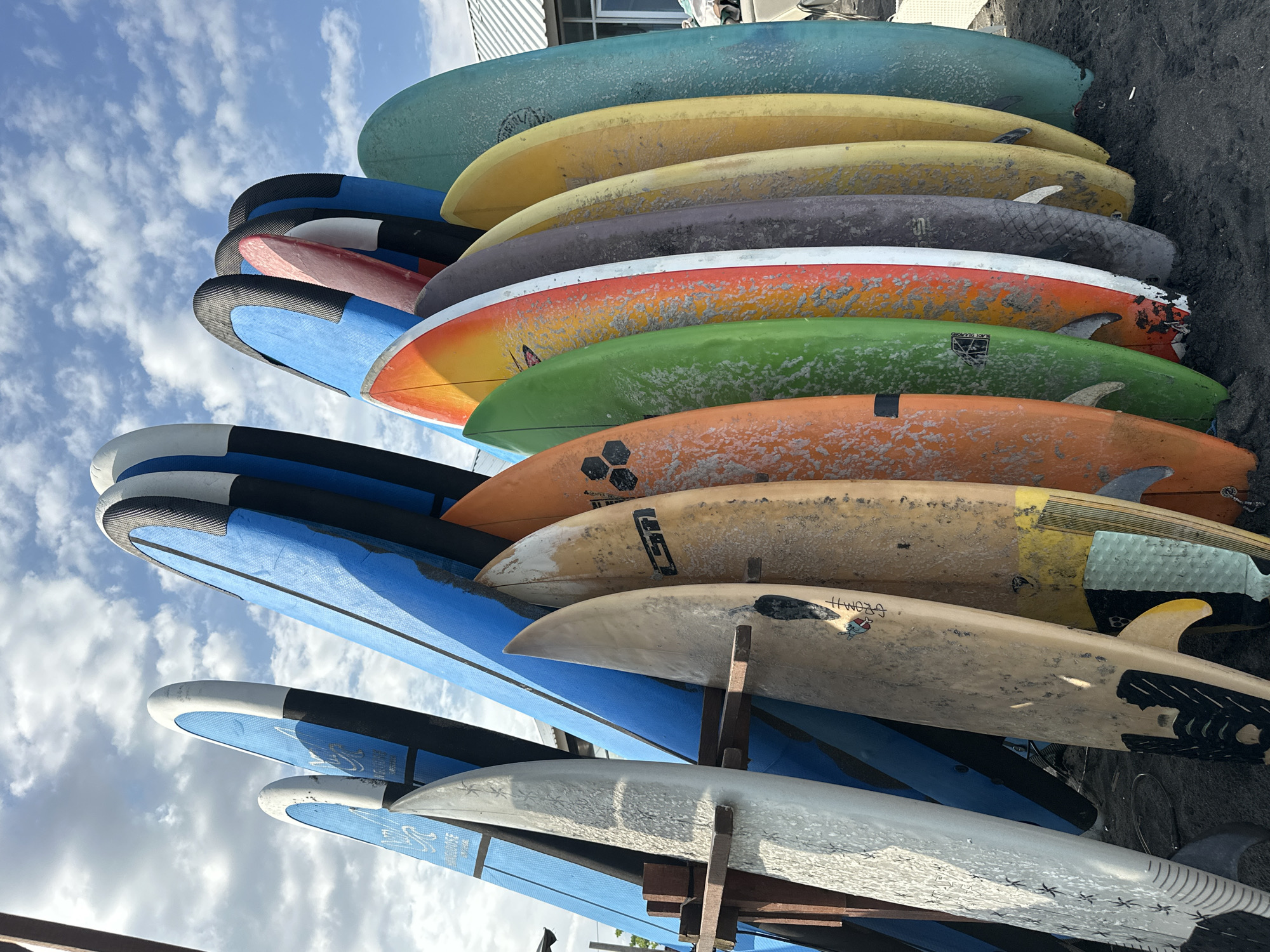 The image shows a collection of surfboards of various colors and designs leaning against a building. The surfboards are stacked in a rack. The sky is visible in the background, with clouds.