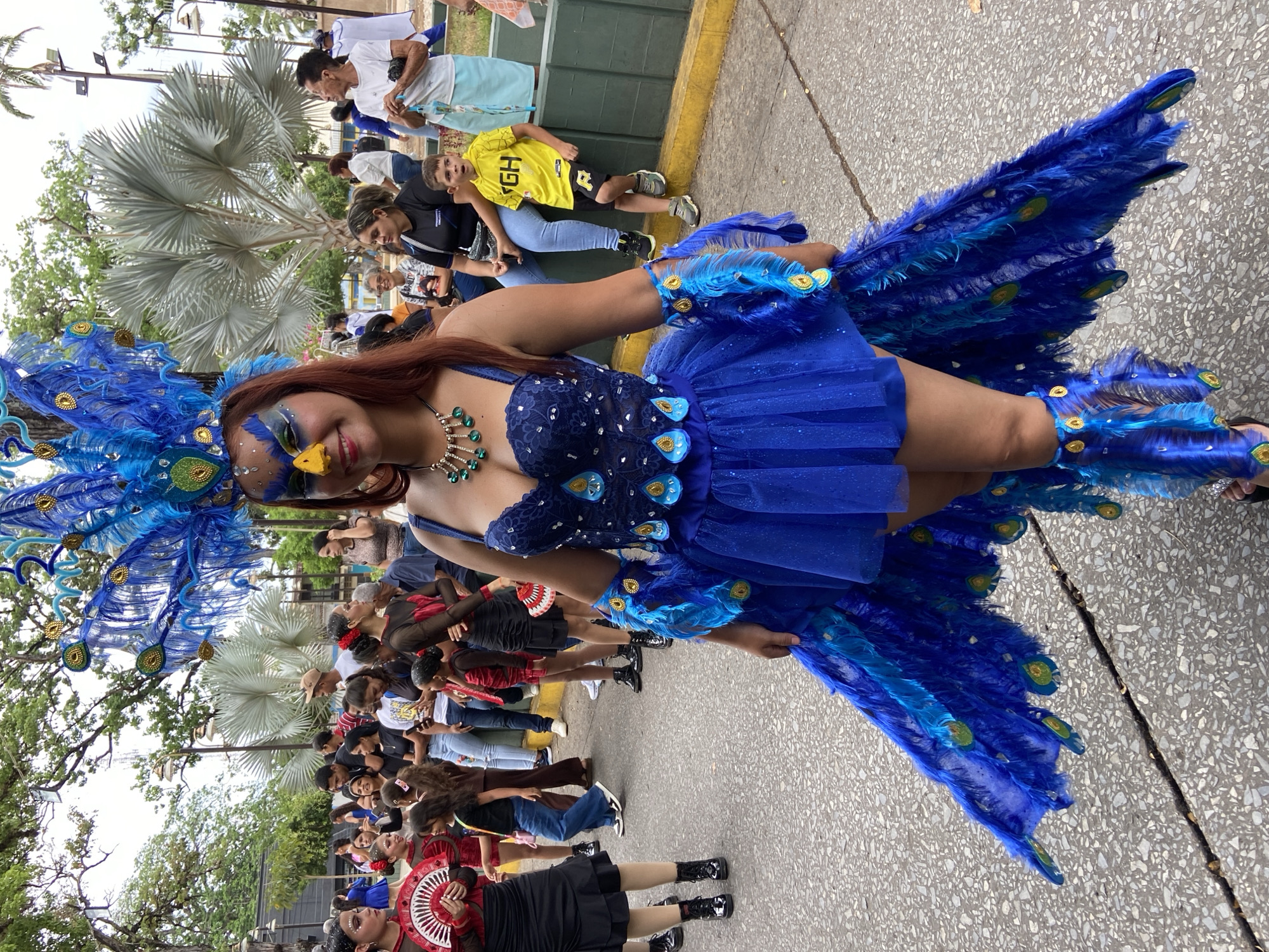 A woman wearing a vibrant blue peacock costume is surrounded by onlookers in Tinaquillo, Venezuela. She has a yellow beak painted on her face. The crowd is a mix of adults and children, some taking photos. The backdrop includes palm trees and a building with green paint. The surface the woman stands on is textured concrete.