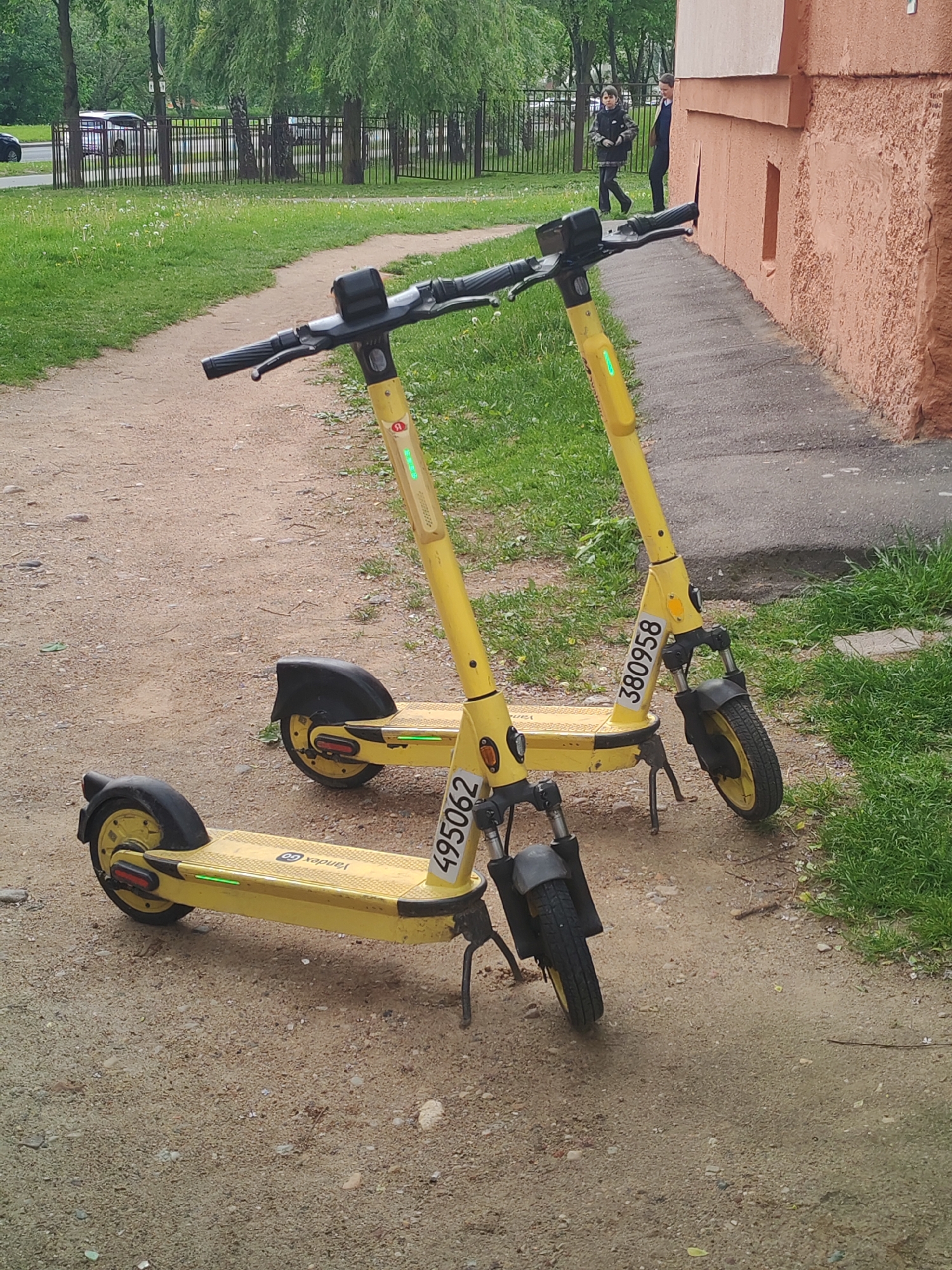 The image shows two parked yellow Yandex Go electric scooters on a dirt path next to a building. The scooter in the foreground has the number "495062" on its side, while the scooter in the background has the number "380958." A person can be seen walking in the background, along with a patch of green grass, trees, and a fence. The location appears to be urban or suburban.