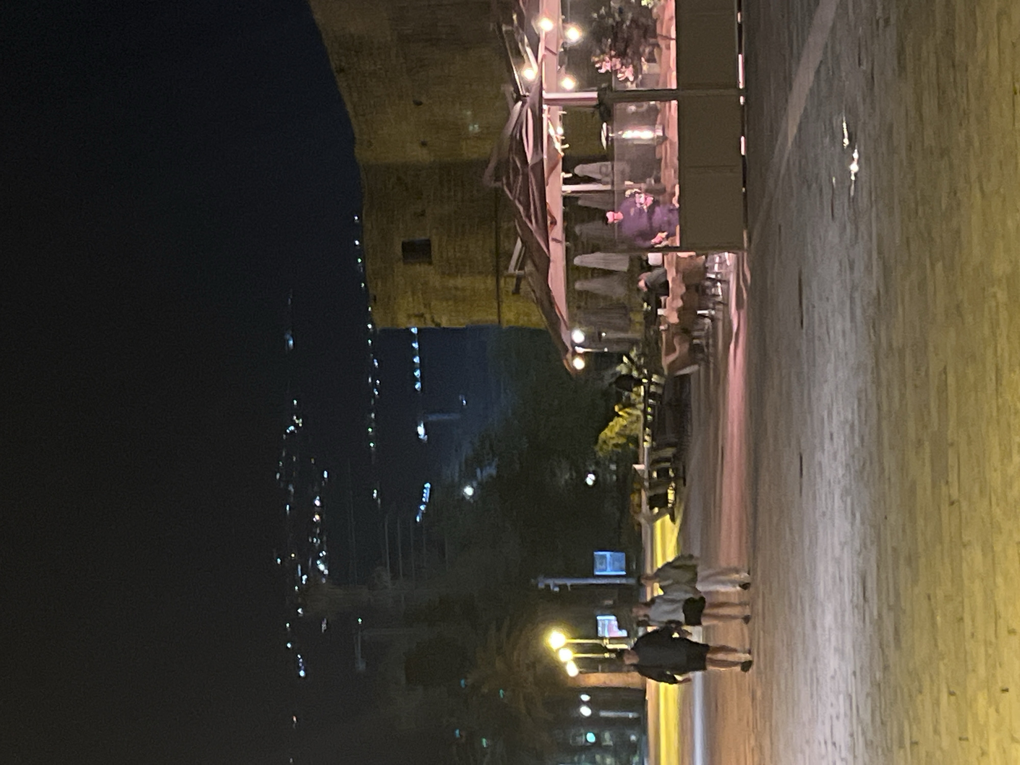 Here is a description of the image:
The image shows a nighttime view of Budva, Montenegro. In the foreground, there is a paved walkway where three people are walking; one appears to be a man in a dark jacket and shorts. Adjacent to the walkway is a restaurant with outdoor seating; people are visible dining. Behind the restaurant is an old stone building. The background features the dark outline of mountains and a view of the lights of the city across the bay.