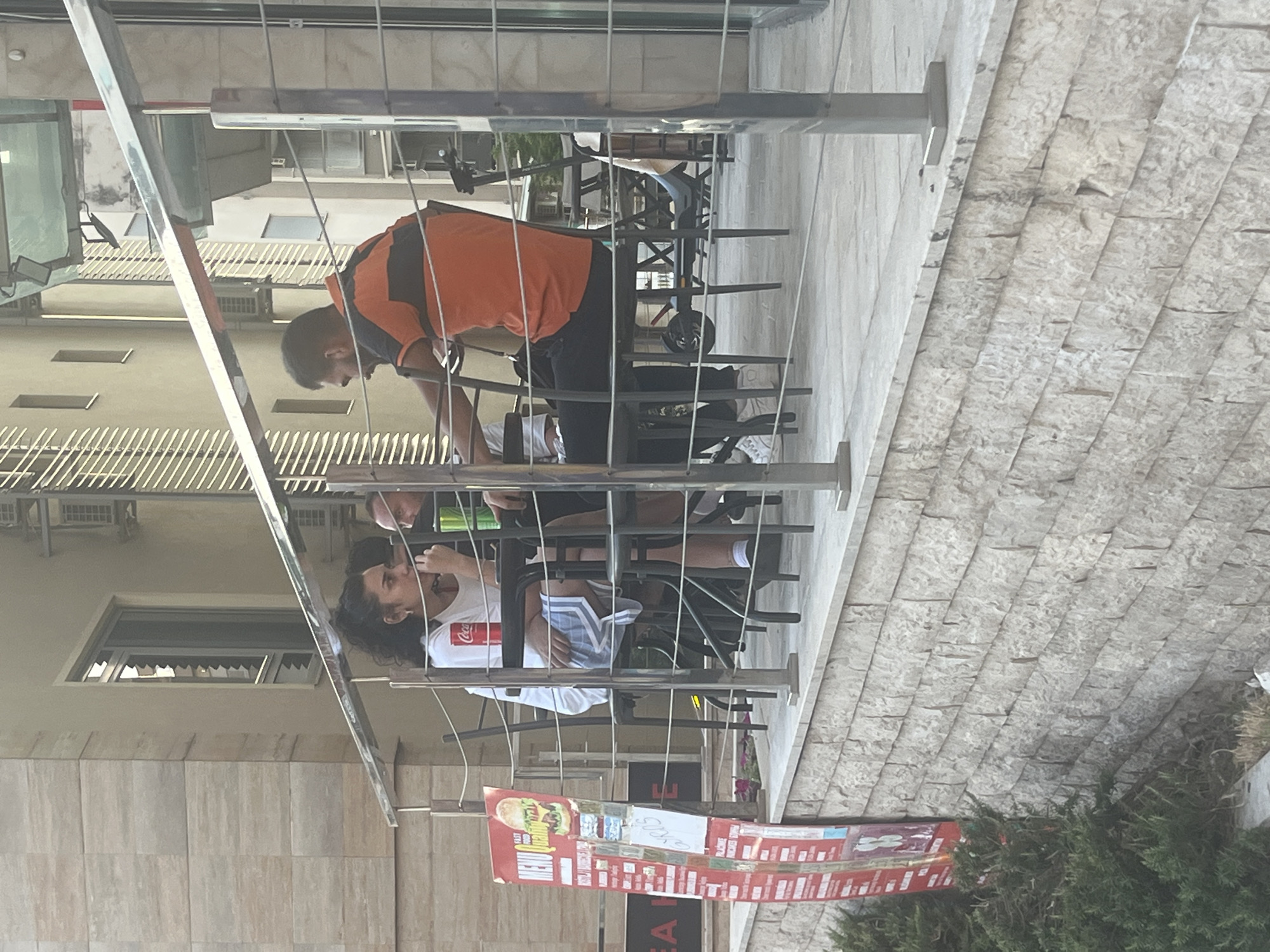 Two individuals are seated at a table on an outdoor patio, which appears to be part of a restaurant or cafe, given the presence of a menu board. The woman, on the left, is wearing a white t-shirt and shorts. She is holding a drink. The man, on the right, is wearing an orange and black striped shirt and dark pants. He is looking towards the woman. A Coca-Cola can is visible on the table between them. The patio is bordered by a railing and overlooks a street or walkway. The building behind them is modern, with light-colored facades and windows. There is also a prominent menu board to the left of the table, displaying food items and prices. An electric scooter is parked beneath the table on the right. The overall scene is casual and appears to be set during the daytime.
