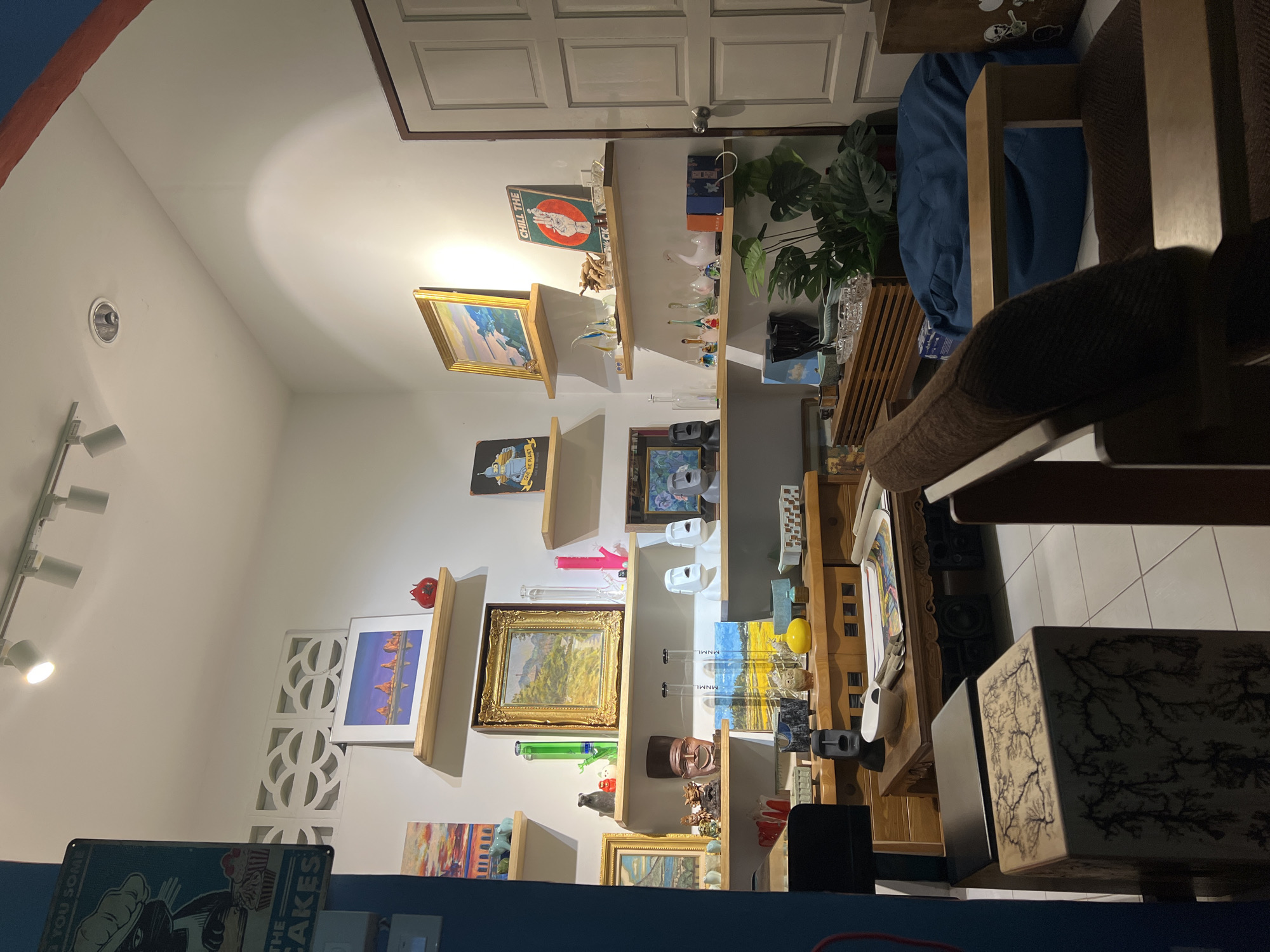The image presents an interior scene featuring a white wall adorned with numerous shelves holding various items such as paintings, figurines, and decorative objects. A track lighting system illuminates the wall from the left side. To the right, the scene transitions into a more cluttered area with furniture, including a chair and a table. The orientation of the objects suggests the image has been rotated. The location is Unknown, Unknown, and there are no people visible.