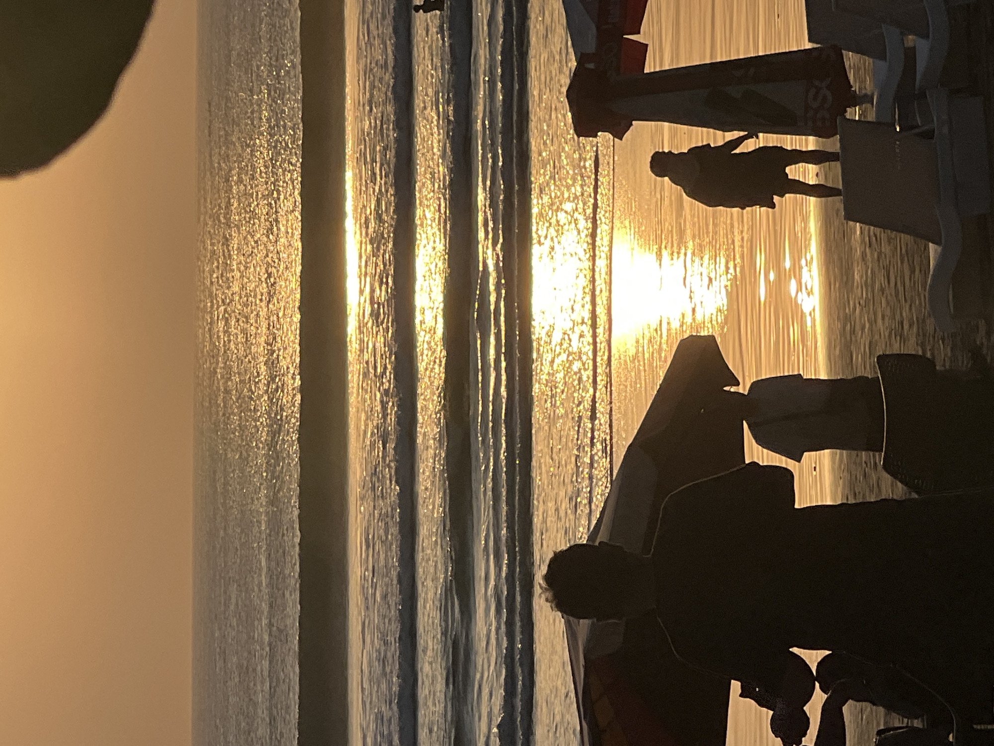 The image depicts a beach scene at sunset on Kuta Beach, Indonesia. The sky is a warm orange hue, and the setting sun casts a shimmering golden reflection on the ocean waves. In the foreground, silhouetted figures are visible, some standing and others appearing to be seated on lounge chairs. Two umbrellas stand on the beach, their shapes casting shadows. The ocean has several rows of gentle waves rolling towards the shore. Further out in the water, a few people are visible, some wading and one person appearing to be swimming. The overall impression is one of a serene and tranquil evening at the beach.