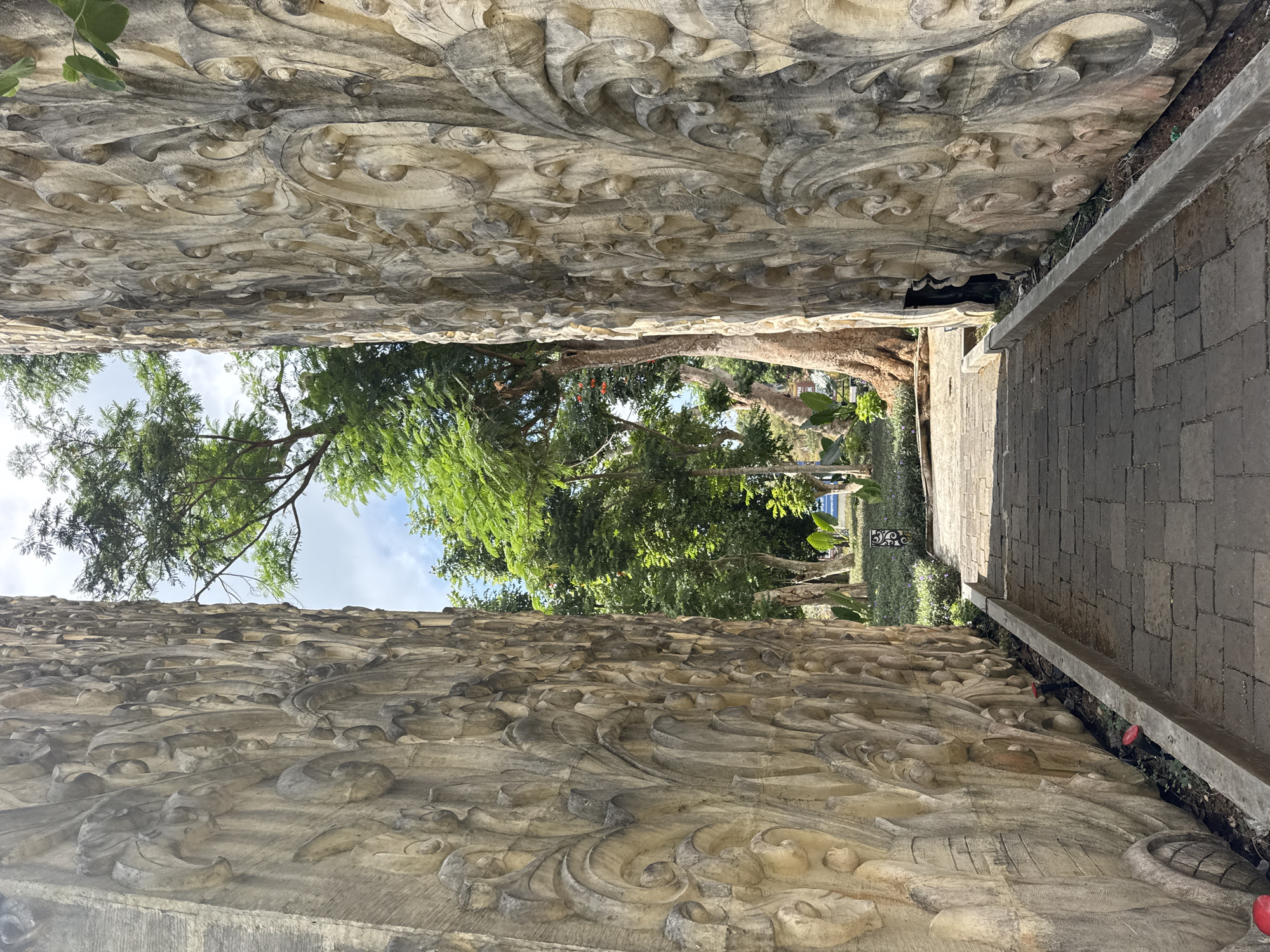 The image shows a pathway leading through a narrow opening between two large, intricately carved stone walls. The walls are adorned with repetitive, ornate patterns that appear to be floral or abstract in nature, rendered in a beige or light brown stone. The pathway is paved with rectangular stone tiles and is bordered by a concrete curb. Beyond the walls, a lush green environment is visible, with mature trees, foliage, and a partly cloudy blue sky. The pathway appears to be in an outdoor public space, likely a garden or park. In the distance, hints of further vegetation and possibly a building can be seen. The overall impression is one of an artistic and natural setting. The provided location is Kuta Selatan, Indonesia.