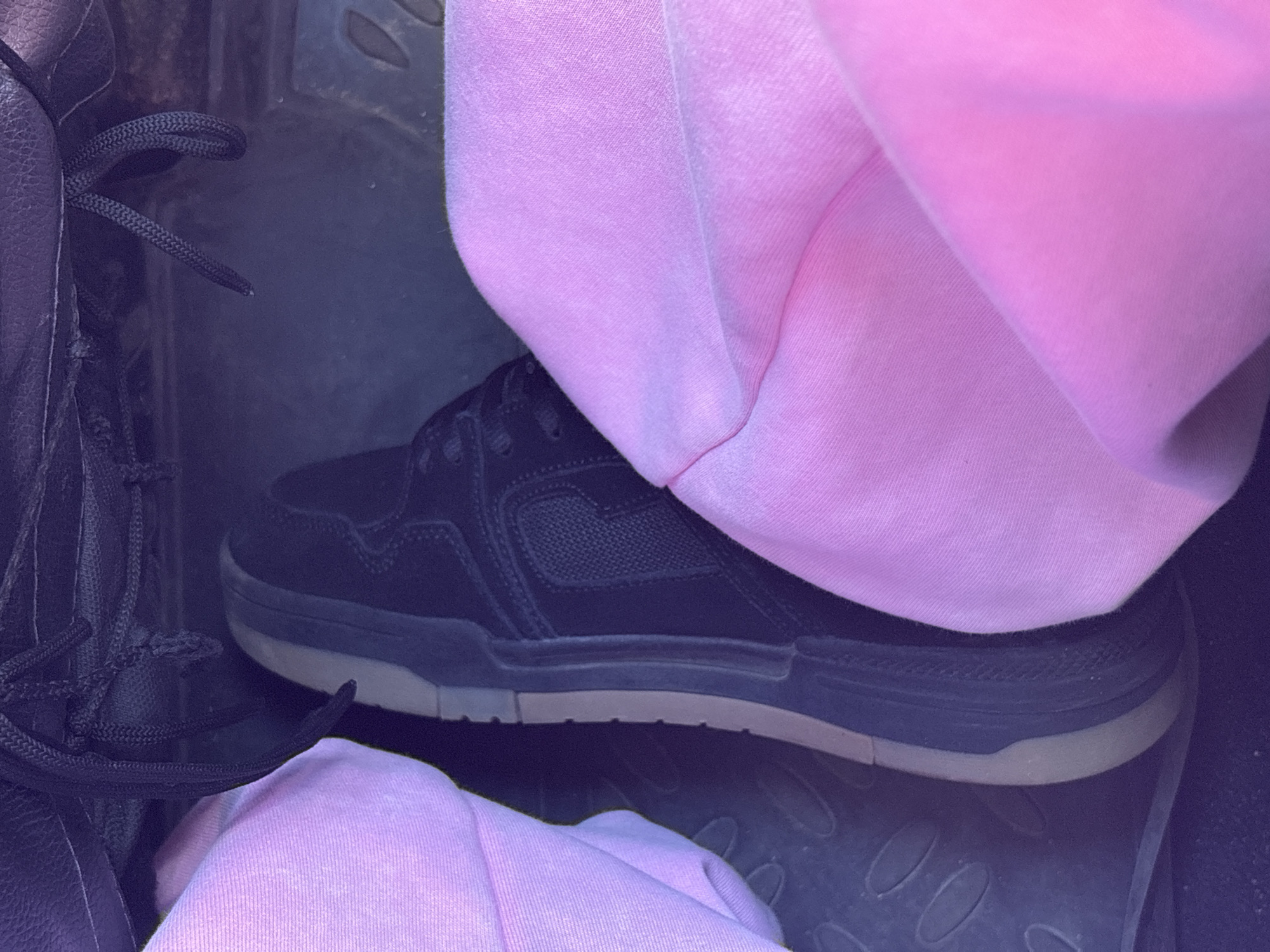 The image shows a close-up view of a person's feet in a vehicle. The person is wearing black sneakers with light brown soles and light pink sweatpants. The sneakers are resting on the car's floor mats, which have a textured pattern. The background is dark and out of focus, suggesting the interior of a car. The lighting is dim, indicating it could be daytime with overcast conditions or nighttime. There is no visible text in the image. The scene is simple and focuses on the footwear and clothing.