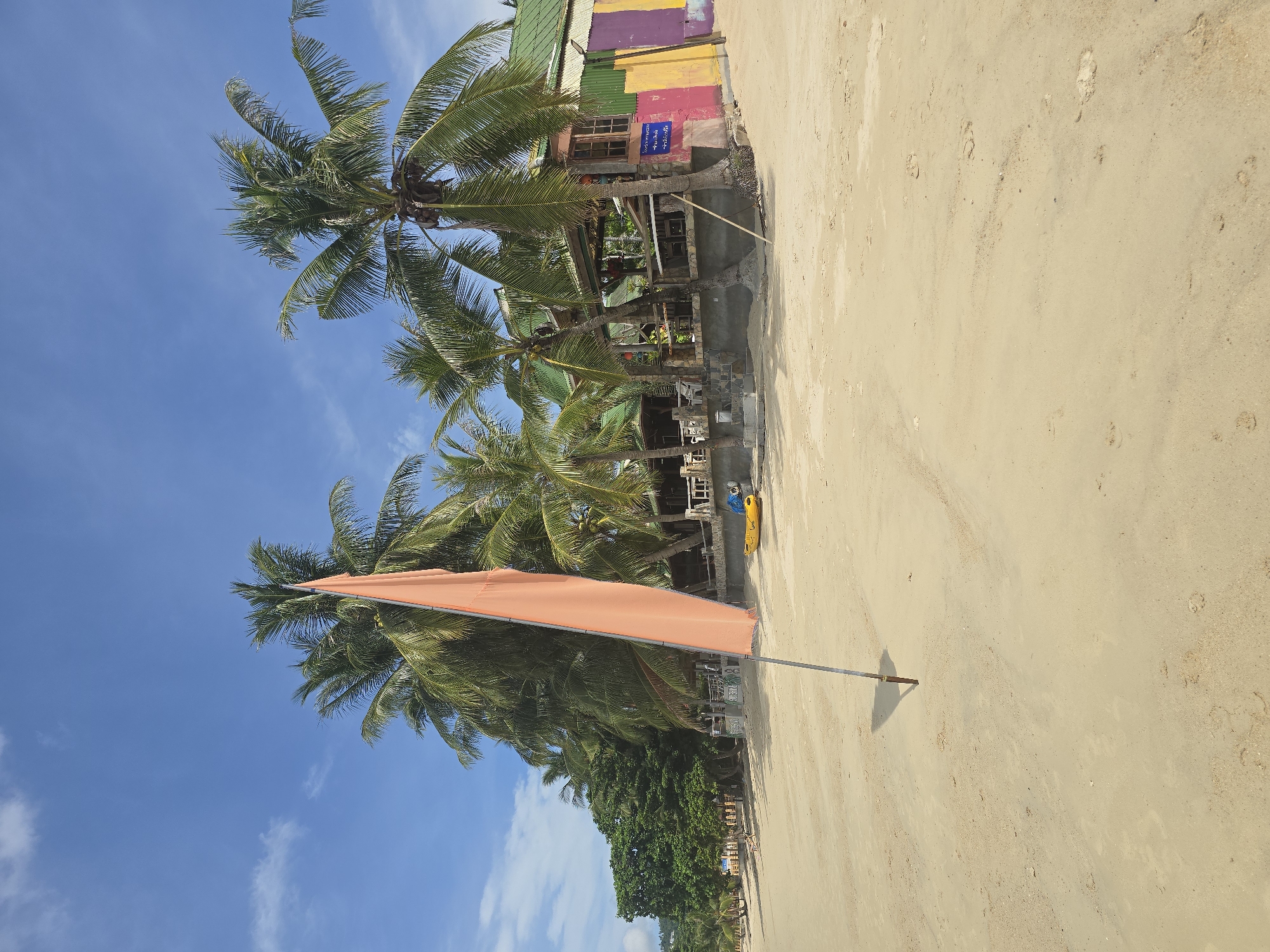 The image depicts a beach scene on a sunny day in Baan Lamai, Thailand. In the foreground, a sandy beach stretches out with footprints visible in the sand. A single orange flag on a pole stands prominently in the middle ground, casting a shadow. Behind the flag, a line of palm trees borders the beach. Further back, a colorful building with a green roof is visible, appearing to be a restaurant or bar, with tables and chairs set up. A yellow kayak is resting on the sand near the building. The sky is blue with scattered white clouds.