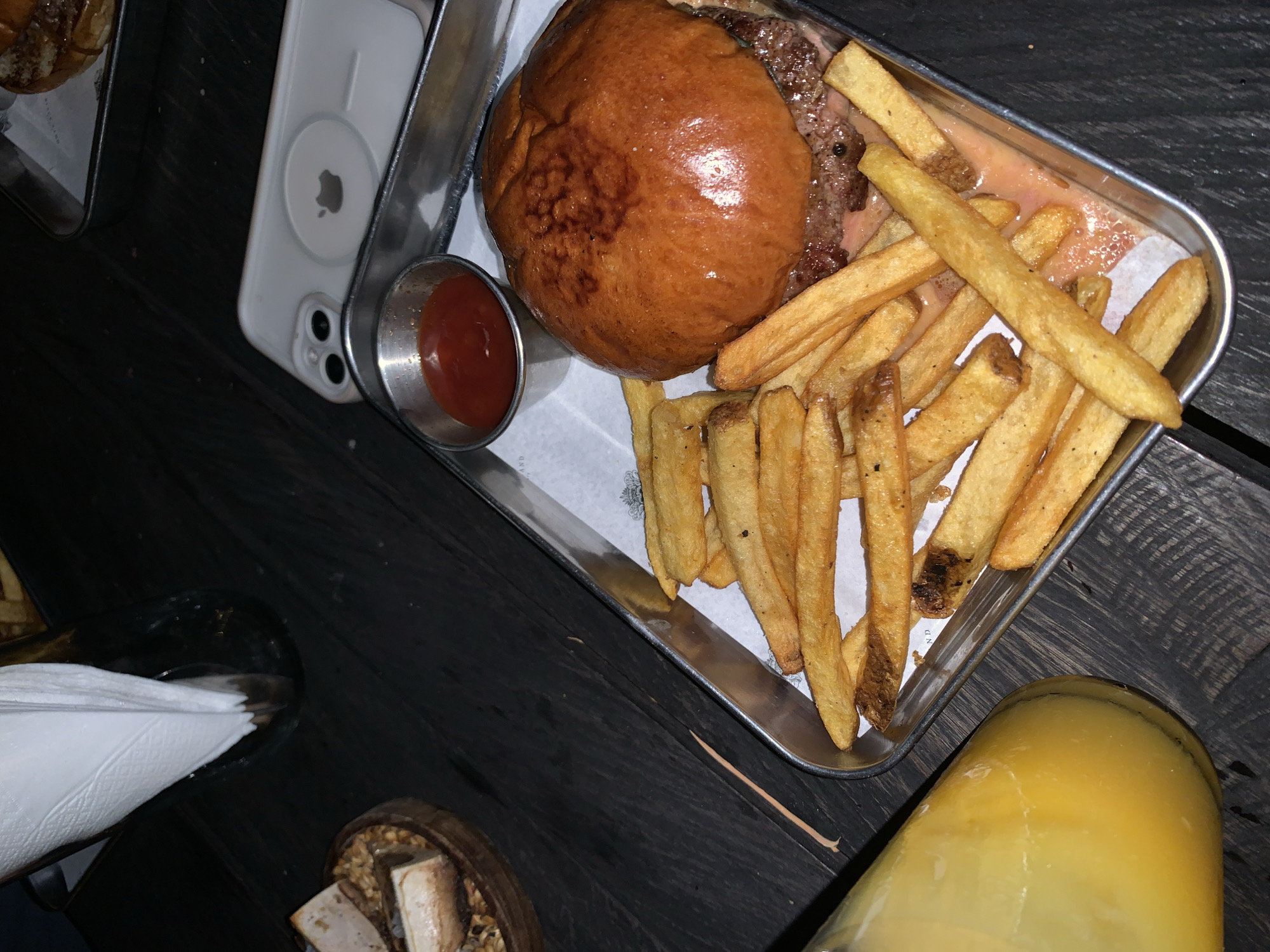 The image shows a metal tray containing a burger and fries, along with a small container of sauce, sitting on a dark wooden table. Next to the tray is a white smartphone with a visible camera module. A glass containing what appears to be juice and a napkin holder with napkins are also partially visible. The scene is set indoors, possibly in a restaurant or bar. The lighting is dim. I cannot confirm that the location is in North Kuta, Indonesia.