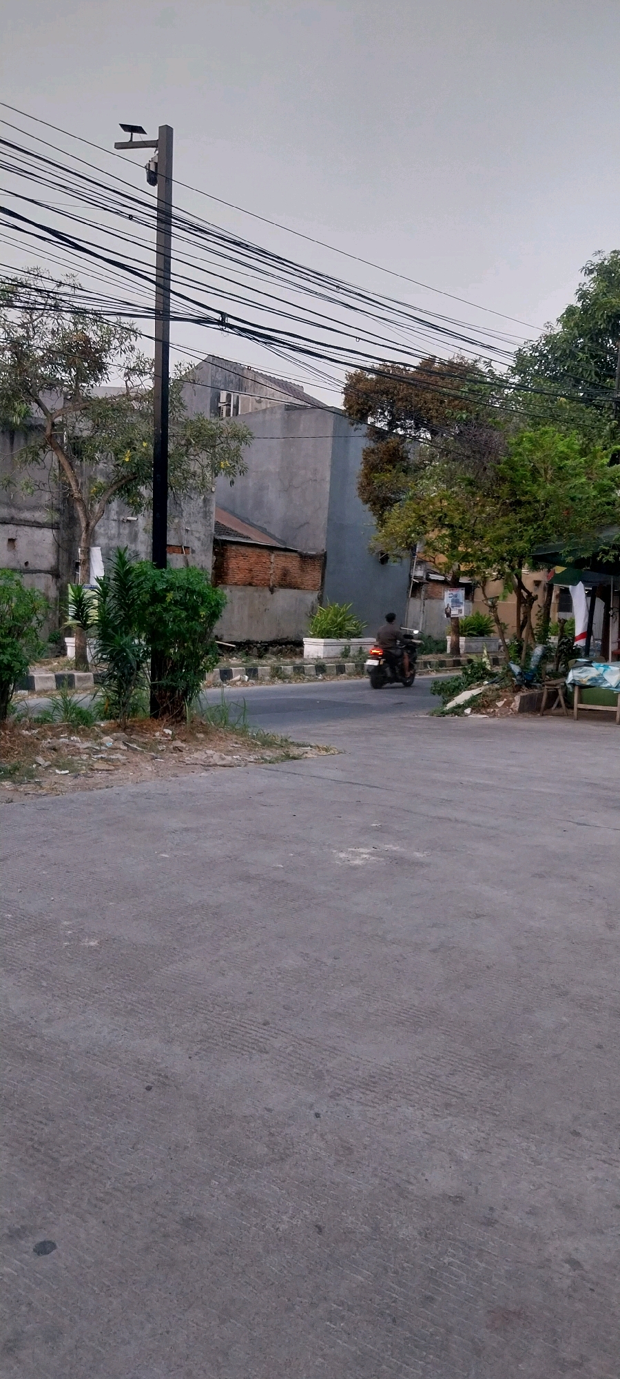 The image captures a street scene in Bojong Nangka, Indonesia. In the foreground, a gray asphalt road dominates the lower half of the frame. In the midground, a single person is riding a dark-colored motorcycle away from the camera, heading towards the right side of the image. The motorcycle has its taillight illuminated. To the left of the road, there are several green bushes and a tree with a gray trunk. Further back, concrete buildings with gray walls and some exposed brickwork are visible. A tall, dark utility pole with numerous black wires stretching across it stands prominently on the left side of the image. The sky is overcast, suggesting a cloudy day. On the right side of the midground, there appears to be a small roadside stall or structure with some items displayed.