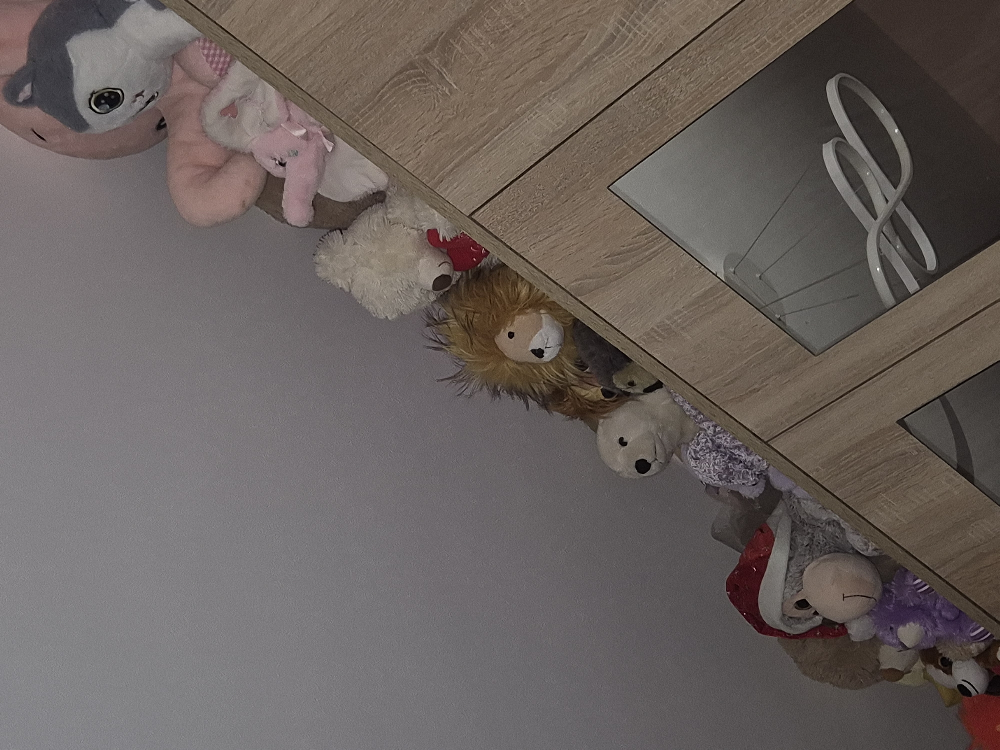 A collection of stuffed animals is displayed on an angled wooden shelf unit. The stuffed animals are of various sizes and types, including a large pink elephant, a grey and white cat, a light-colored bear holding a red heart, a lion, a white bear, a monkey wearing a Santa hat, and a purple stuffed animal. The shelf unit has glass doors on the lower section, revealing a modern light fixture inside. The background is a plain grey wall. The image is taken from a slightly low angle, looking upwards.