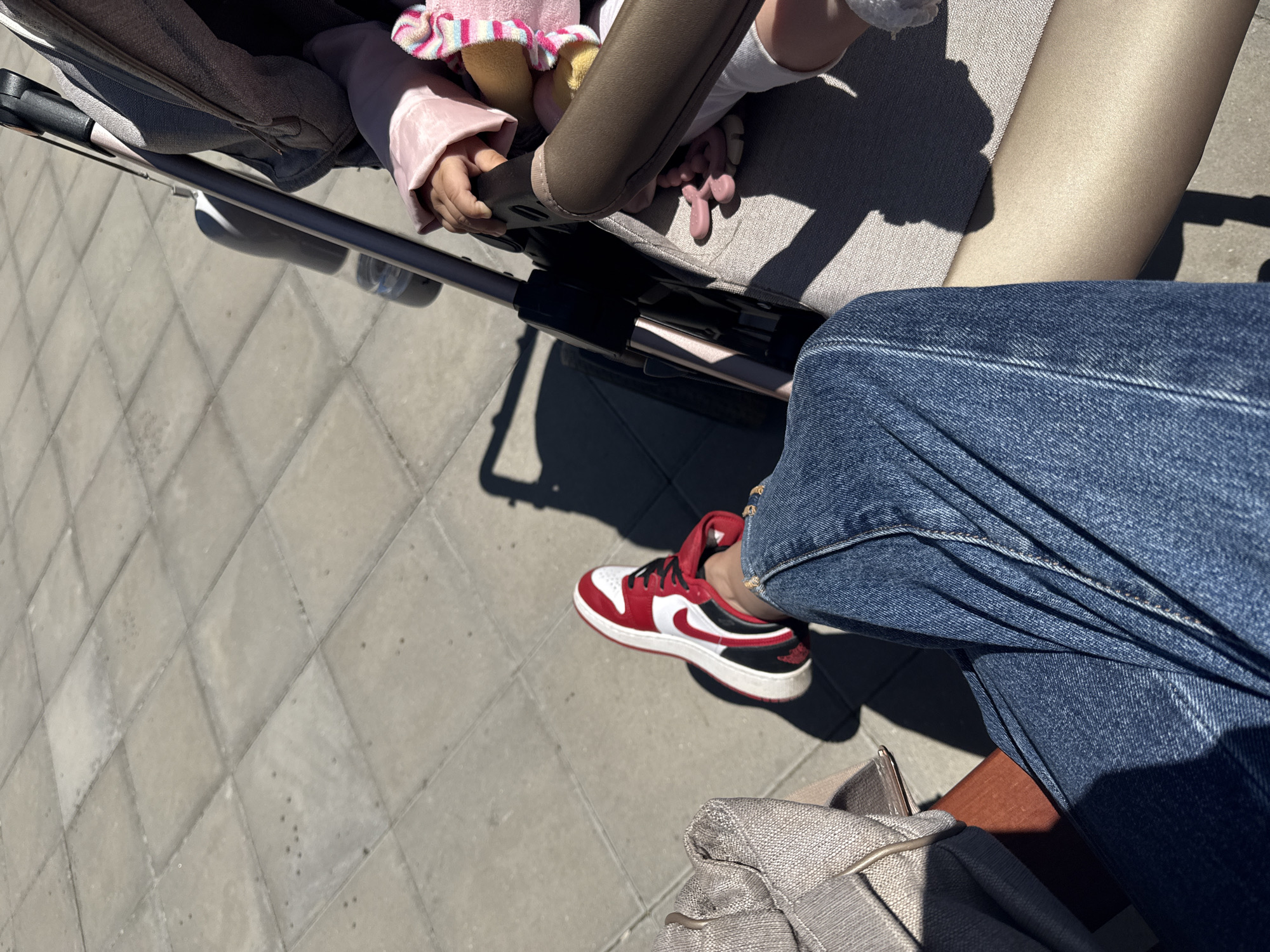 The image captures a stroller on a paved surface, presumably a sidewalk or pedestrian area, in an unidentified urban location. A child is visible in the stroller. The person pushing the stroller is wearing blue jeans and red, white, and black sneakers. A bag or purse is also present. The time of day seems to be during daylight hours.