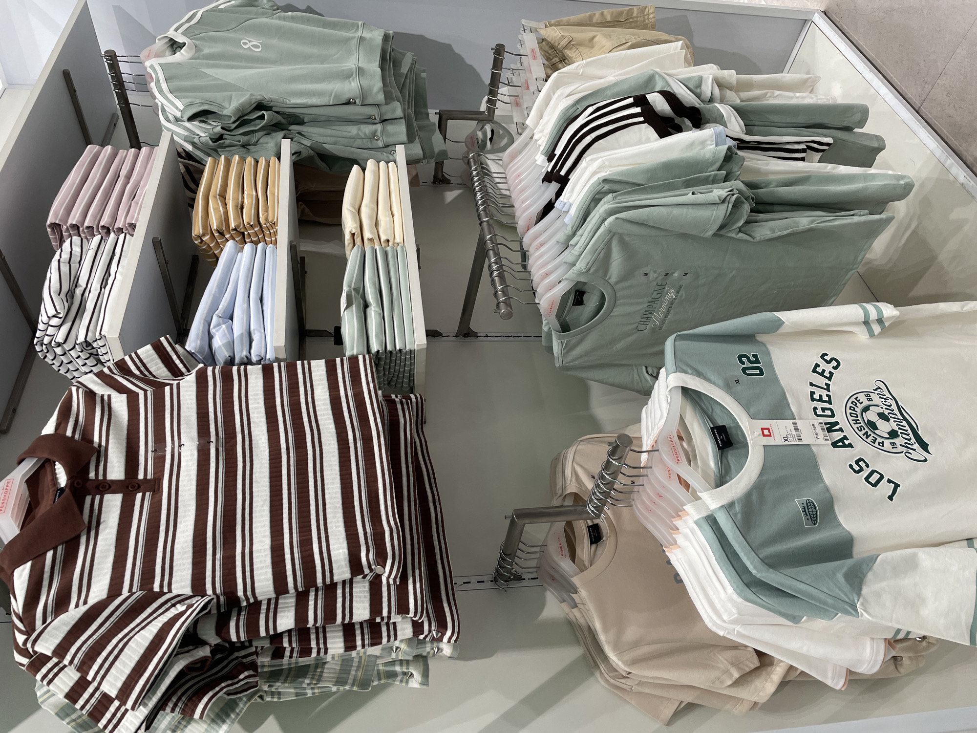 A clothing store display in Davao City, Philippines, showcases an array of apparel on tiered shelves and hanging racks. The topmost shelf displays folded tops in various patterns and colors, including horizontal stripes of brown and white, and solid pastel hues. Below this, more folded tops are visible, in light blue and tan.
On the right side, a rack holds several light green long-sleeved tops, some featuring white stripes on the sleeves and the number "80" printed on the chest.
The lower section of the display features hanging t-shirts on white and pink hangers. These include plain tan shirts, white shirts, and t-shirts with a color-block design in white and light green, one of which has graphics of "LOS ANGELES Champions" and a soccer ball. Another t-shirt in light green has the text "CHAMPAGNE Morning". The background is a plain light-colored wall. No people are visible in the image.