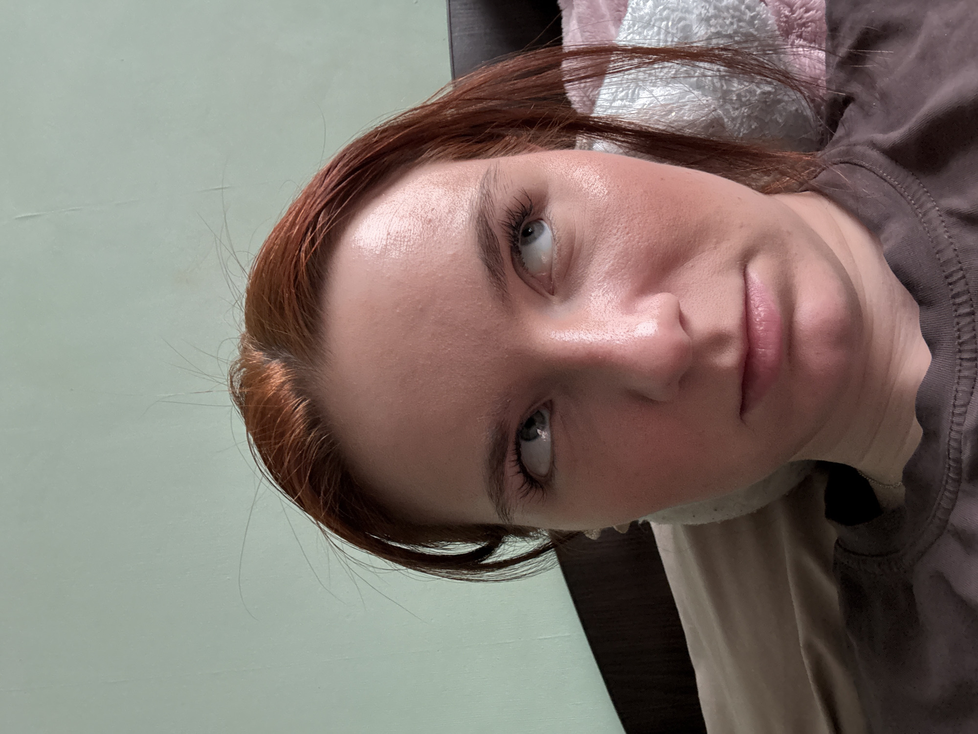 A red-haired person is lying down. The background shows a light green wall and other soft materials in neutral colors. The person appears to be wearing a brown t-shirt. The city and country of the image are not discernible from the provided visual information.