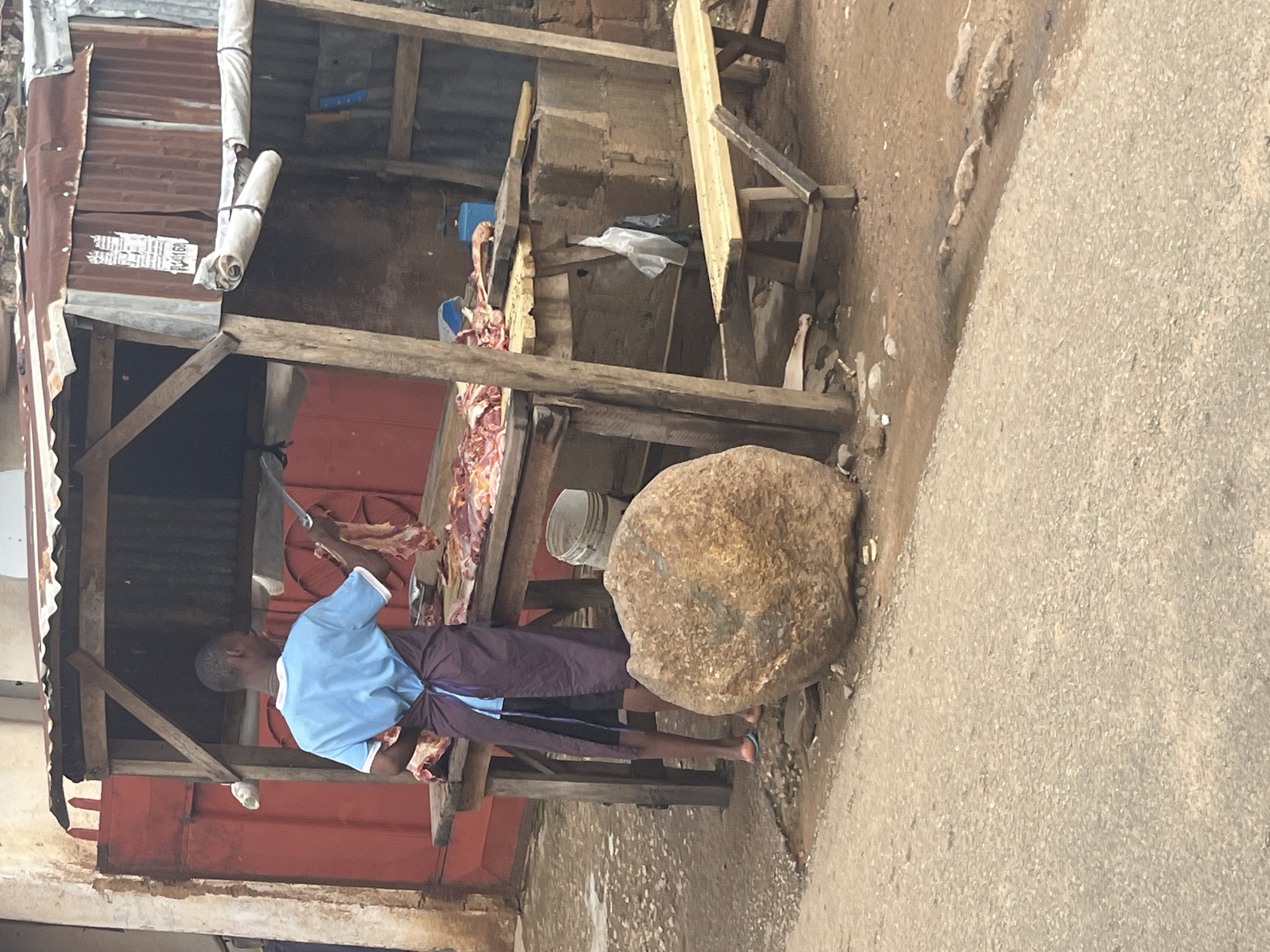 The image depicts an outdoor butcher's stall, likely located in Jos, Nigeria, given the context provided. A young Black male, wearing a light blue t-shirt and a dark purple apron over black shorts, stands behind a wooden table displaying cuts of raw meat. He is holding a large knife in his right hand and appears to be in the process of preparing or cutting meat. The stall itself is constructed from rough wooden beams and corrugated metal sheeting, suggesting a basic, open-air setup. Behind the butcher, more raw meat is visible on the table, along with what appears to be a scale and a blue plastic container. To the left of the stall, a large, round, brown boulder is prominently positioned. In the foreground, a grey, asphalt road is visible, and the ground surrounding the stall is a mixture of dirt, gravel, and some debris. The overall impression is a scene of everyday commerce in a bustling urban environment, characterized by its rustic and practical nature.