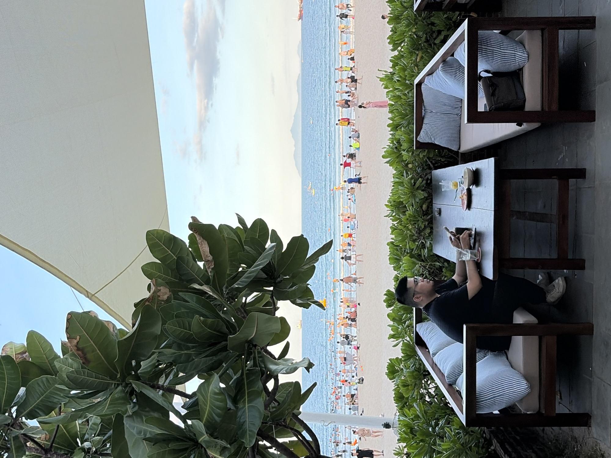 The image depicts a man sitting at a wooden table on a patio overlooking a crowded beach in Đà Nẵng, Vietnam. The man, wearing a black t-shirt and glasses, is looking at his phone. On the table are a glass of juice, a plate with food, and a phone. He is seated on a couch with striped cushions. In the background, the beach is filled with people swimming, sunbathing, and walking. The ocean stretches to the horizon, with several boats visible. Lush green foliage frames the foreground, with a large tree on the left and a hedge separating the patio from the beach. The sky is a clear blue with a few scattered clouds. The overall scene suggests a relaxed, resort-like atmosphere on a sunny day.