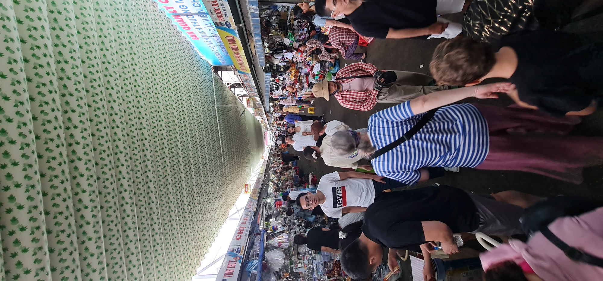 The image shows a crowded marketplace in Đà Nẵng, Vietnam. Numerous people walk through the narrow aisles lined with vendors displaying goods. The vendors are selling various items, possibly including clothing, household goods, and food. Many shoppers are examining the merchandise. The scene is covered by a striped ceiling. Storefront signs in Vietnamese are visible.