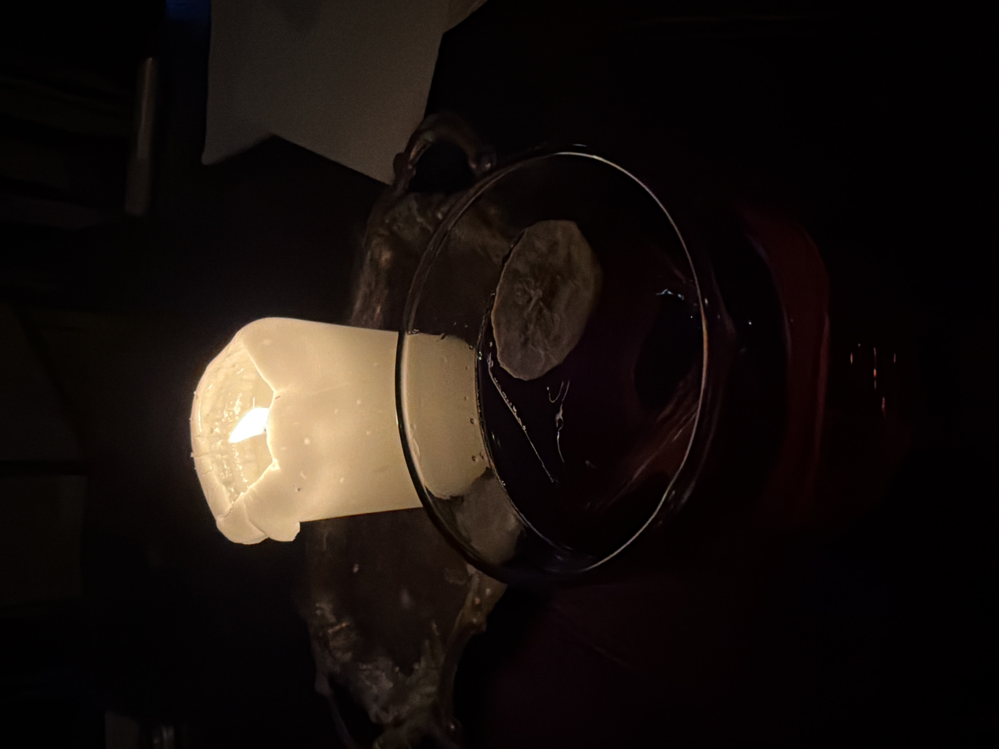 A dimly lit interior features a lit, ivory-colored candle on a tarnished silver tray or holder. Next to the candle, a clear wine glass partially filled with a dark red liquid and a slice of fruit (likely lemon or lime) sits on a dark surface. The surroundings are dark and partially obscured. The image was taken in Tbilisi, Georgia.