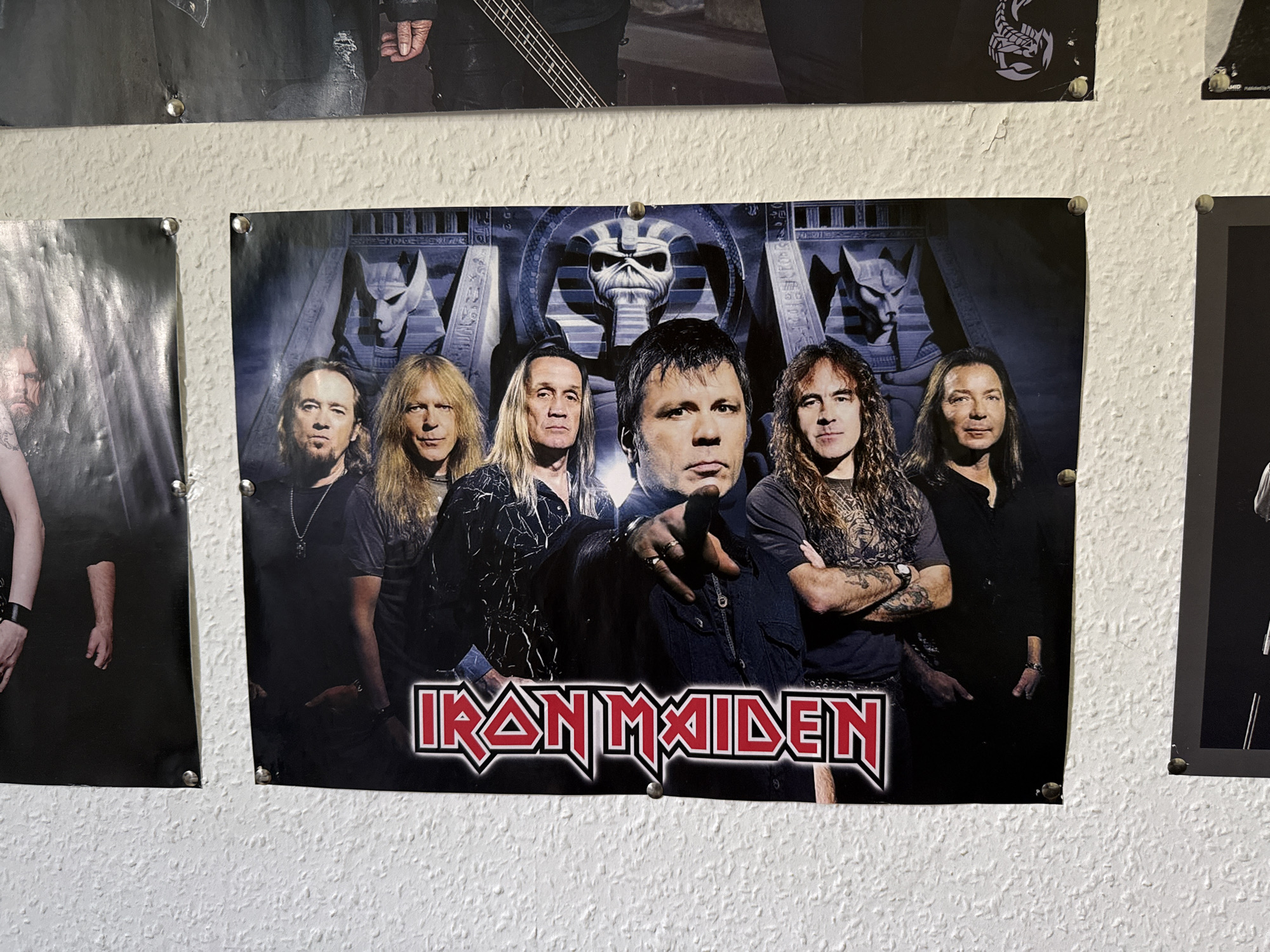 The image depicts a wall in the town of Bad Sachsa, Germany, adorned with several posters. The central and most prominent poster features the band Iron Maiden, with the band members posing in front of a backdrop depicting Egyptian-themed imagery, including a large skull at the top and sphinx-like statues. The band's logo, "IRON MAIDEN," is prominently displayed at the bottom of the poster in a stylized red and white font. To the left of the main poster, partially visible, is another poster featuring a band member from the shoulders down, wearing black clothing and a black wristband. To the right, another poster is partially visible, showing a band member's upper body in black attire. The posters are attached to a textured, off-white wall using visible thumbtacks. The lighting suggests an indoor environment.