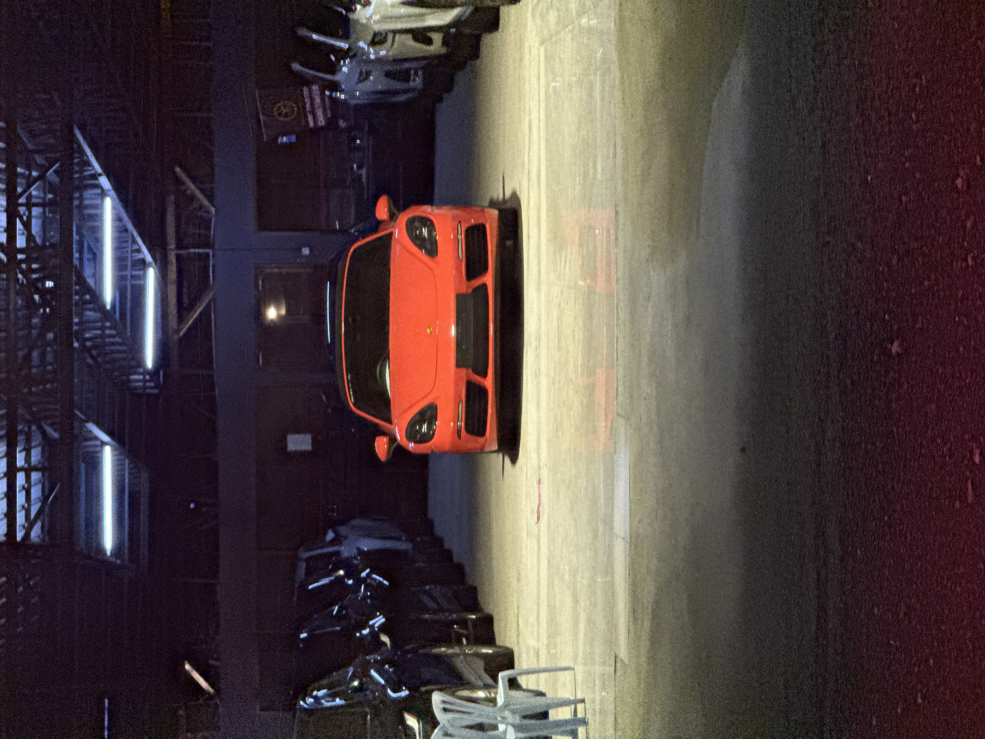 The image depicts an indoor scene, likely a car showroom or a large private garage, viewed from a slightly elevated perspective. The primary subject is a vibrant red Ferrari sports car, parked prominently in the center foreground and facing away from the viewer. Its sleek lines, distinctive front grille, and headlights are clearly visible.
The setting is characterized by low, artificial lighting, primarily from long fluorescent tubes mounted on an industrial-style ceiling structure that spans the upper left portion of the image. This lighting casts shadows and highlights, creating a dramatic atmosphere. The floor is made of light-colored concrete or a similar material.
In the mid-ground, to the left of the red Ferrari, several other cars are parked in rows, mostly dark-colored and less distinct, suggesting a collection or storage area. Some white plastic chairs are also visible in the foreground, near the parked cars.
In the background, on the far left, an architectural framework of beams and possibly stairs or ramps can be seen, partially illuminated by the overhead lights. On a wall in the background, a red sign with white text is visible, which upon closer inspection, clearly reads "FIRE EXIT" and includes a circular hazard or fire safety symbol.
The overall impression is one of a controlled environment, likely evening or night due to the reliance on artificial light. There are no people visible, and no specific activity is taking place beyond the cars being displayed or stored. The image has a slightly grainy texture, indicative of low-light photography. The scene does not contain any specific landmarks identifying Kuala Lumpur, Malaysia, but is consistent with an urban indoor car facility.