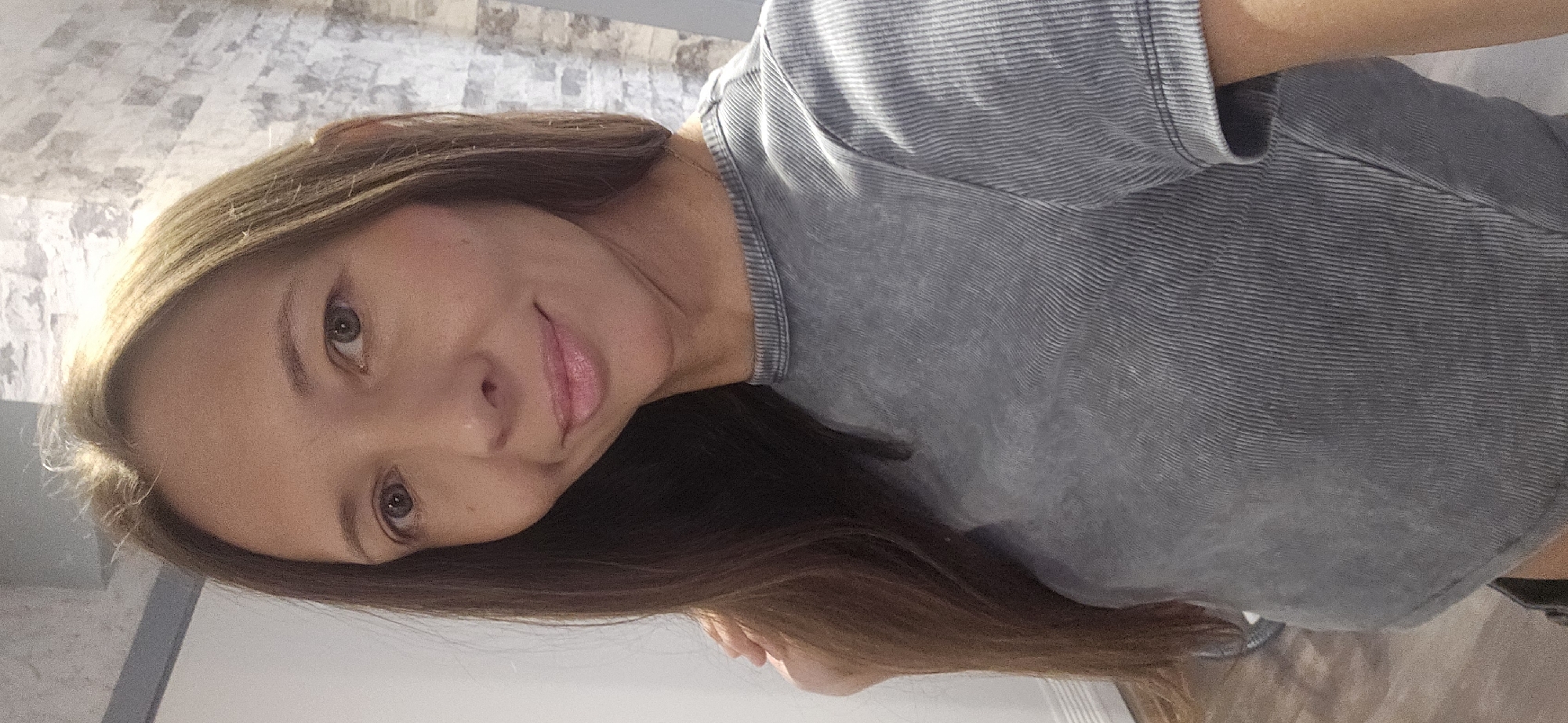 A woman is standing indoors, posing for a selfie in a dimly lit room with a grey, ribbed t-shirt and dark bottoms. The background includes a white wall, a brick wall with grey and white tones, and a wooden floor. The woman has long brown hair and is looking directly at the camera with a slight smile. The lighting suggests an indoor setting, possibly a home, with artificial light reflecting off her hair and face. There is no indication of the city of Vitsebsk, Belarus, in the image.