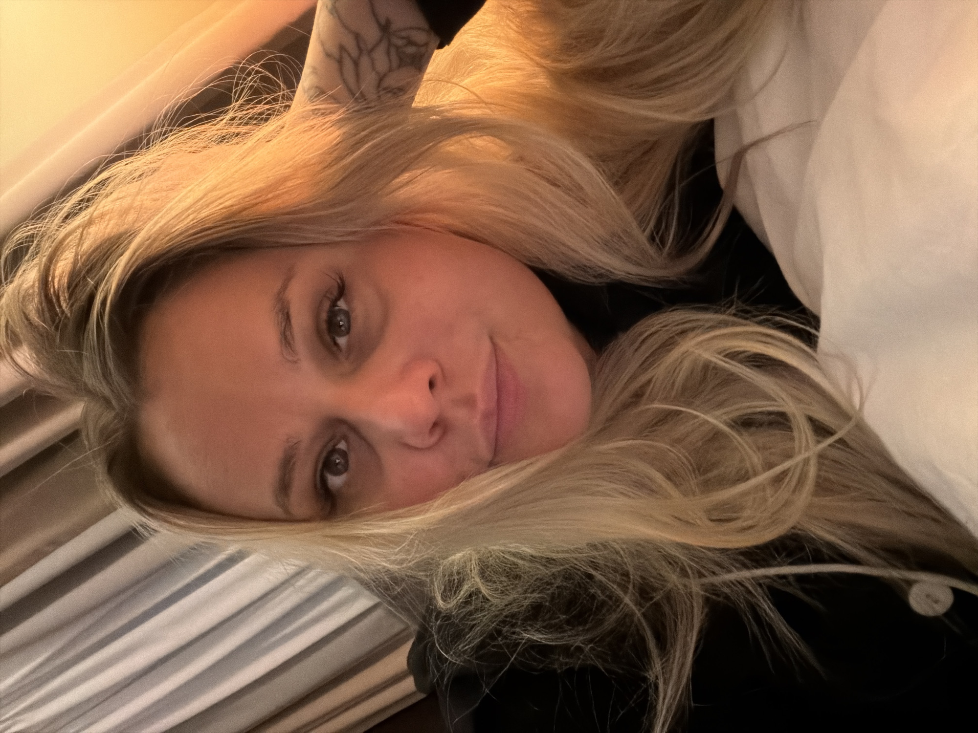 An adult female is depicted in a close-up, likely a selfie, in an indoor setting. She has fair skin, prominent brown eyes, and long, wavy blonde hair with darker roots, which is slightly disheveled and frames her face. Her lips are closed, and her expression is neutral, with direct eye contact towards the camera. She wears a dark-colored garment, possibly a black hoodie or sweater, with a small white circular button or bead visible near her left shoulder. A tattoo with a floral or intricate pattern is visible on her right inner forearm, positioned behind her head. She is reclining against a light-colored pillow or bedding, visible on the bottom right. The background on the left features vertical striped curtains in muted tones of beige and grey. The background on the right, and parts of her hair, are illuminated by a warm, orange-toned light source, suggesting an interior light or possibly a sunset. The specific location within Sarapul, Russia, cannot be determined from the visual information present in the image.