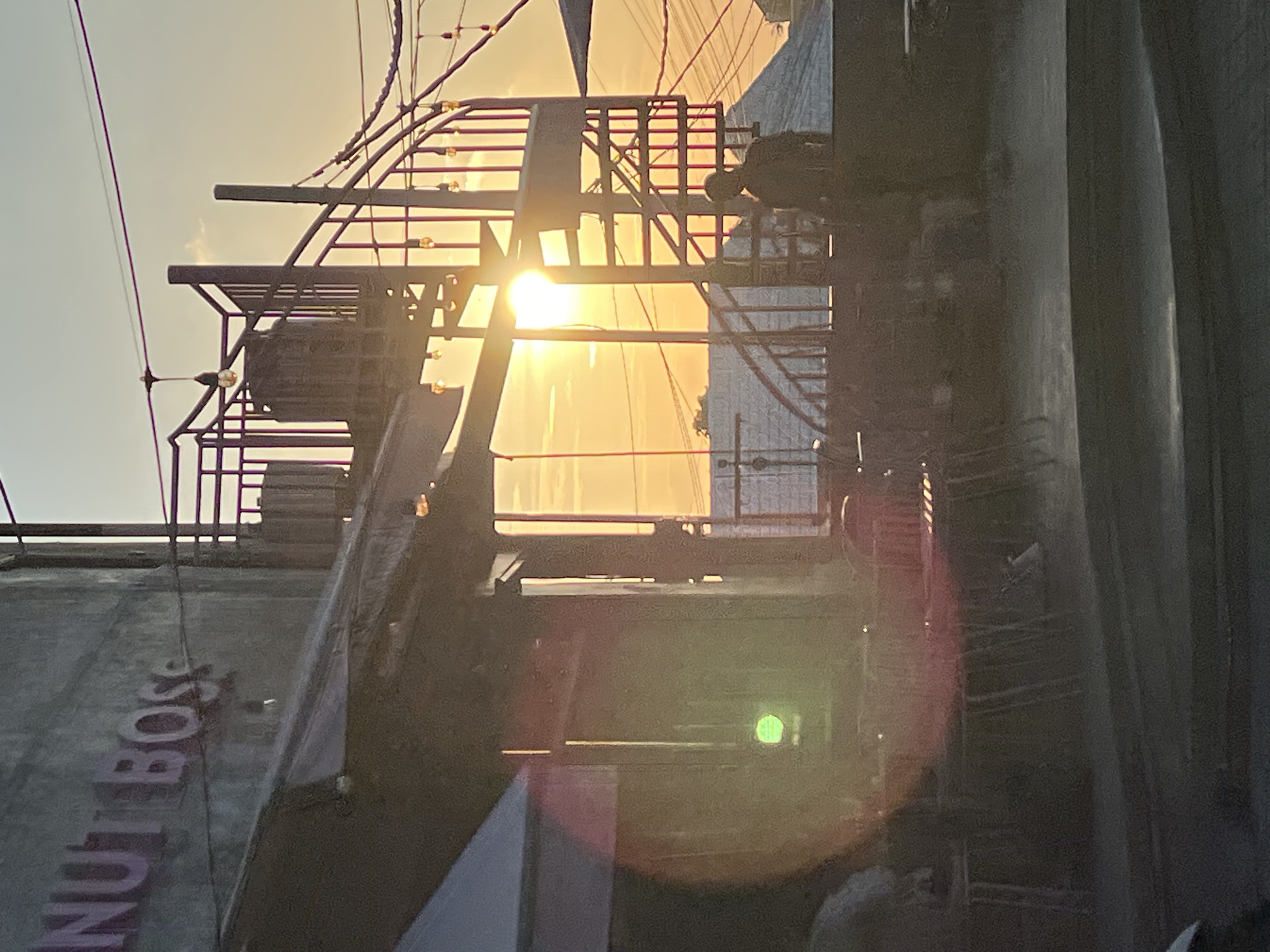 The image shows a building with the word "NUT BOSS" visible on its facade, suggesting it is a commercial establishment. In front of the building, there are tables and chairs, indicating an outdoor seating area. A spiral staircase is visible to the right, leading to an upper level, and it is adorned with string lights. The setting sun casts a warm glow across the scene, creating lens flare in the foreground. The overall atmosphere suggests a relaxed, possibly evening, setting at a restaurant or cafe. This scene is located in North Kuta, Indonesia.