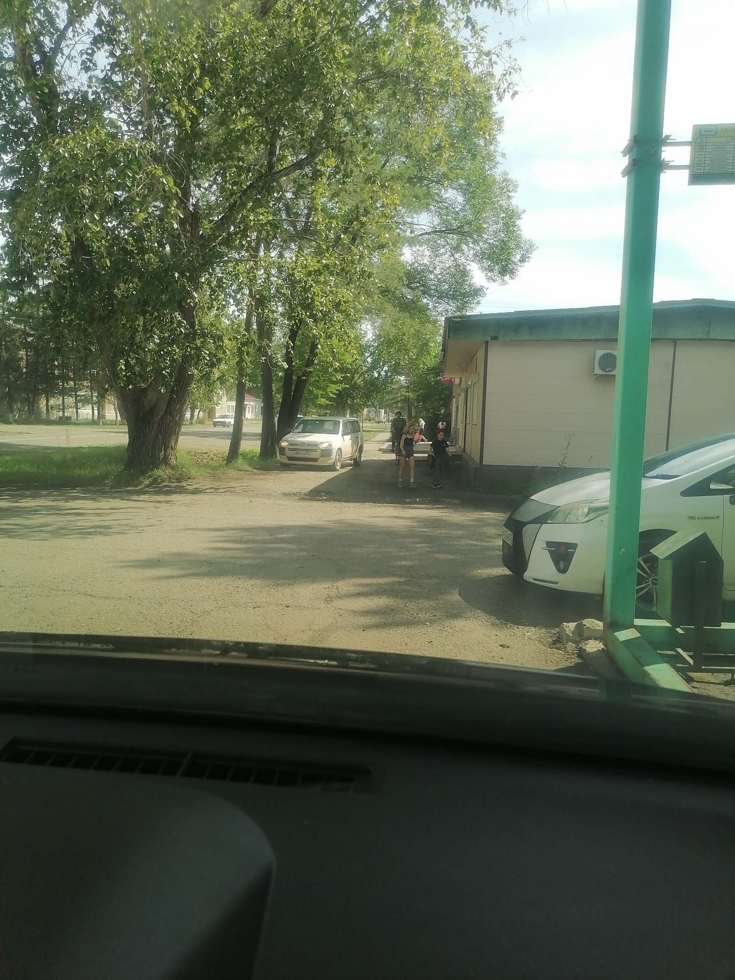 A paved area outside a building in Novopokrovka, Russia, on a bright, sunny day. In the foreground, the dashboard and windshield of a vehicle are visible, suggesting the photo was taken from inside a car. To the left, a large tree with dense green foliage casts shadows on the ground. Further back, a white car is parked on the side of the road, with a building behind it. Two individuals, one male and one female, are standing near the building. The building appears to be a small, beige structure with a green roof. To the right, a green metal pole, possibly part of a bus stop shelter, stands prominently. The sky is a pale blue with scattered clouds.