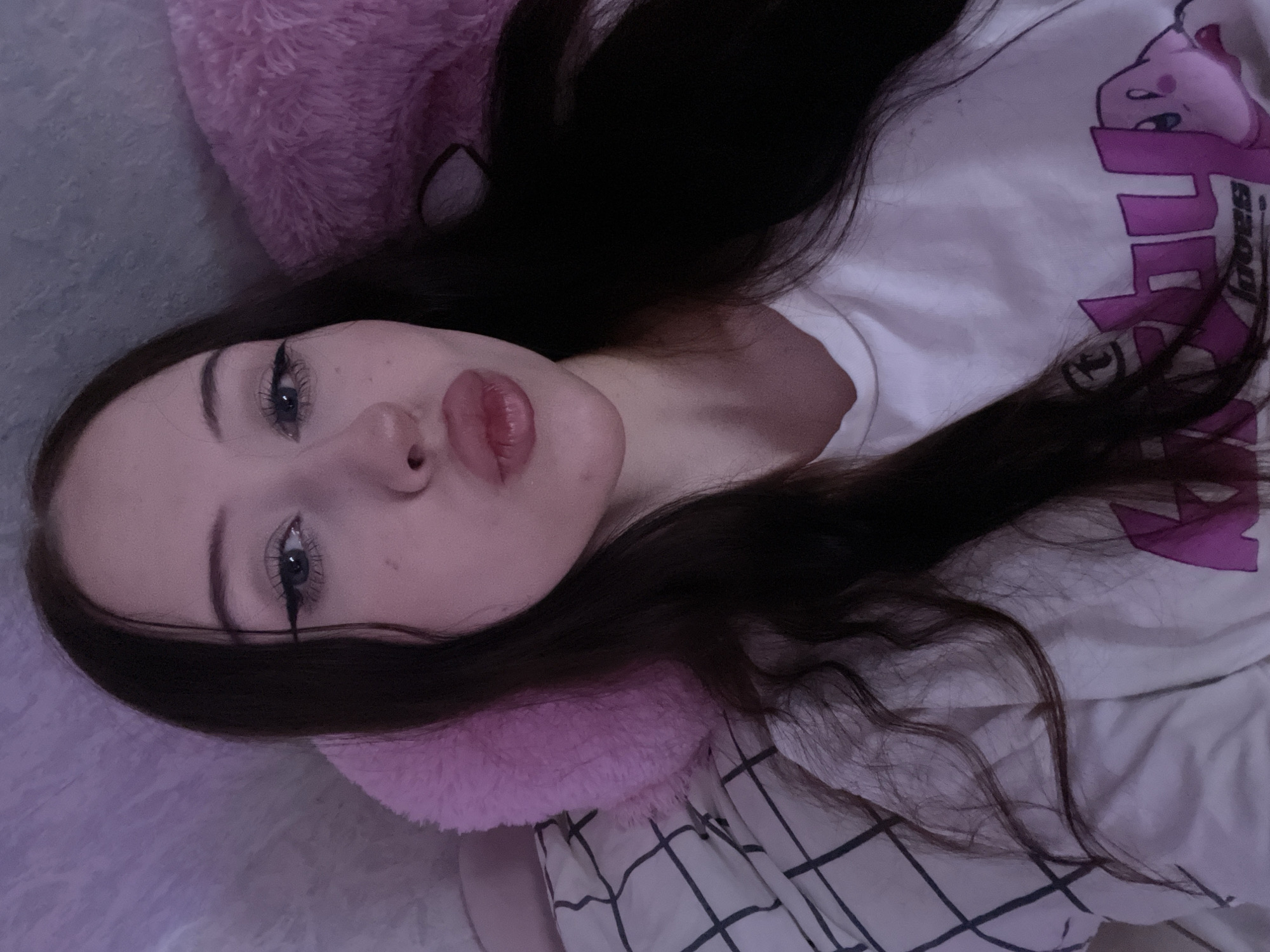 The media file is a close-up, vertically oriented selfie of a young person lying down. The subject has long, dark hair that is spread around their face and shoulders. They are wearing a white t-shirt with a purple graphic print on it, featuring what appears to be a stylized character or logo. The person's face is visible, with makeup applied to their eyes, including eyeliner and mascara, and their lips are slightly parted. They are resting on a soft, pink, furry surface, possibly a pillow or blanket, and a plaid fabric is also visible beneath them. The lighting is dim, suggesting either nighttime or an indoor setting with low illumination. The overall mood appears relaxed and intimate, typical of a casual selfie. There is no visible text in the image, other than what is part of the t-shirt graphic. The location context provided is Votkinsk, Russia.