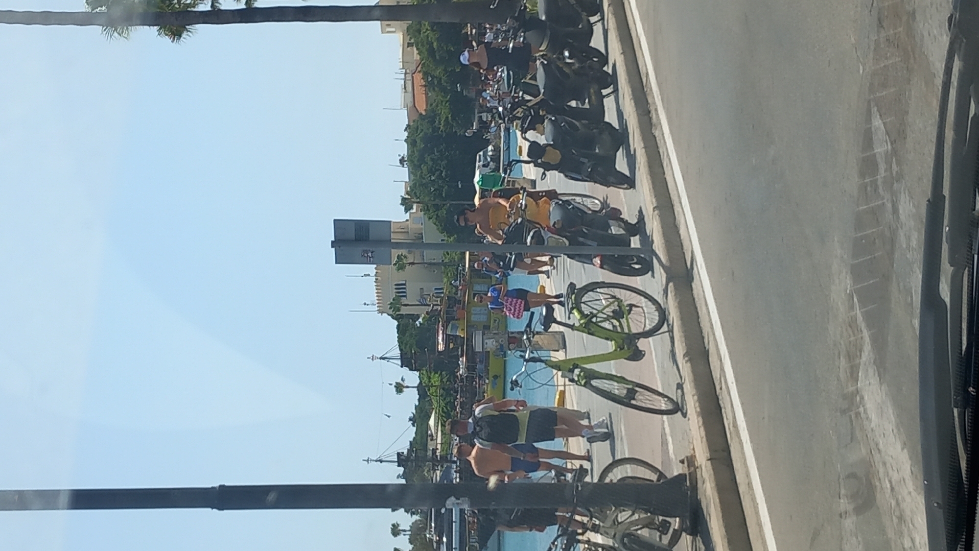 The image depicts a waterfront scene in the city of Kos, Greece, likely near the harbor or a promenade. The foreground shows a street with parked bicycles and scooters. A bright green bicycle is prominent, with a black scooter to its right. In the midground, several people are walking along a pathway adjacent to the water. Some are dressed in casual summer attire, with one woman in an orange top riding a scooter. Behind them, the water features several boats, including a distinctive pirate-themed ship and a yellow boat. In the background, white buildings with arched windows and palm trees suggest a Mediterranean architectural style. The sky is clear and blue. The overall atmosphere is that of a sunny day in a tourist area.