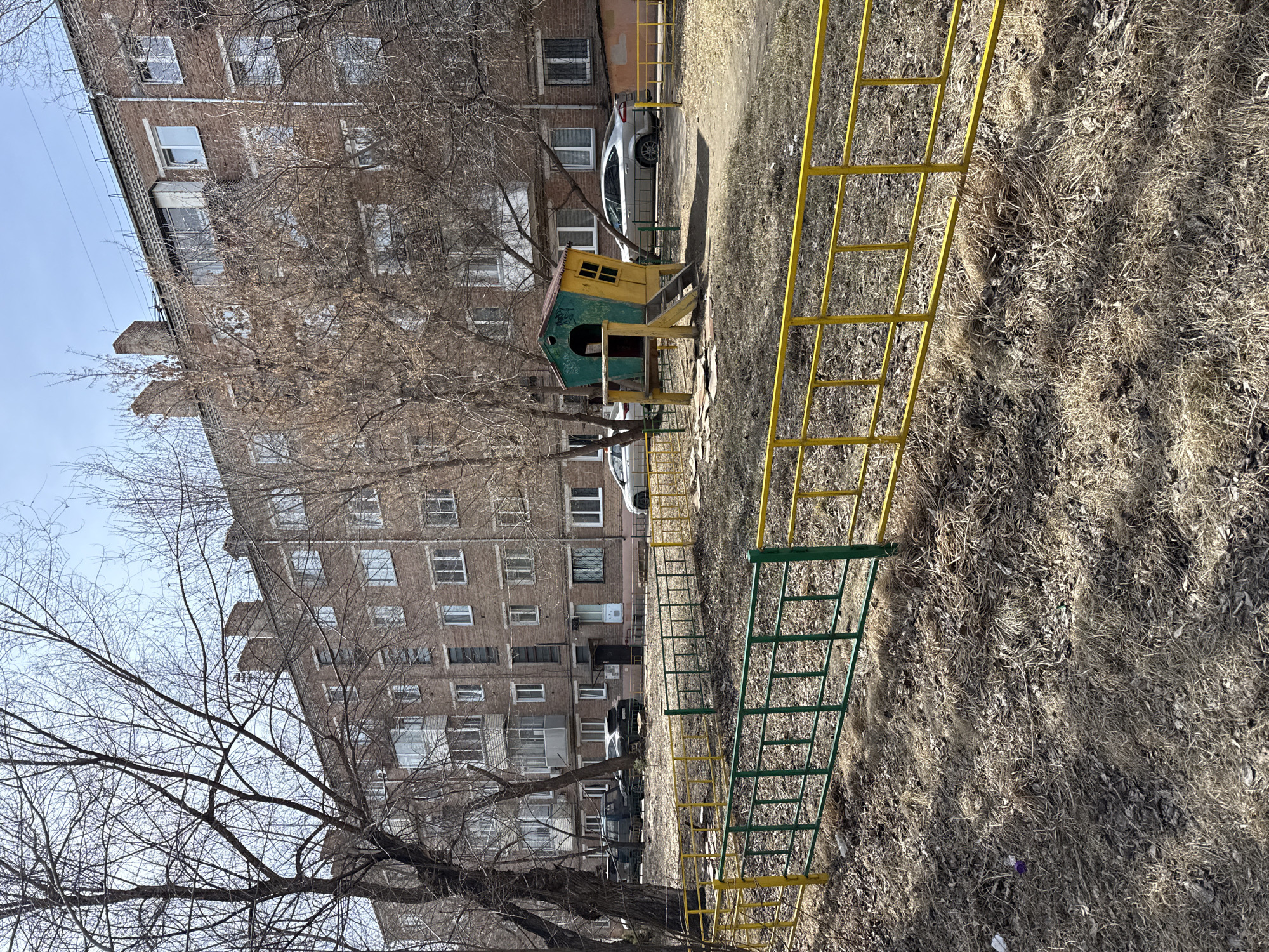 A playground sits adjacent to a brick apartment building on a sunny day. The playground equipment consists of a yellow and green metal climbing structure and a small green and yellow playhouse. Several cars are parked in front of the building. A bare tree stands to the left of the playground. The ground is covered in dry grass and leaves. The photo was taken in Krasnoyarsk, Russia.