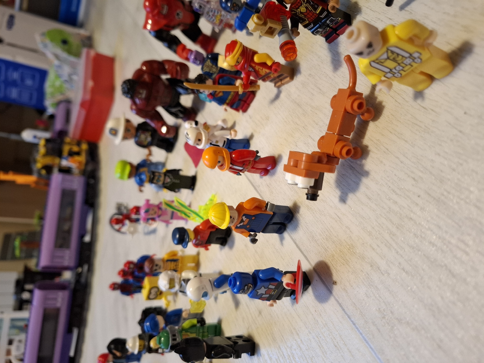 The image depicts a collection of Lego figures and toys scattered on a light-colored, possibly wooden, surface. The figures are diverse, including superheroes like Captain America and what appears to be Hulk, as well as construction workers and other generic characters. A Lego train, predominantly purple, is visible in the background. A small Lego dachshund is positioned in the foreground. The scene is indoors, likely a domestic setting, with toys and a blurry background suggesting a room. There is no indication or contextual information linking this image to the city of Tyumen, Russia.