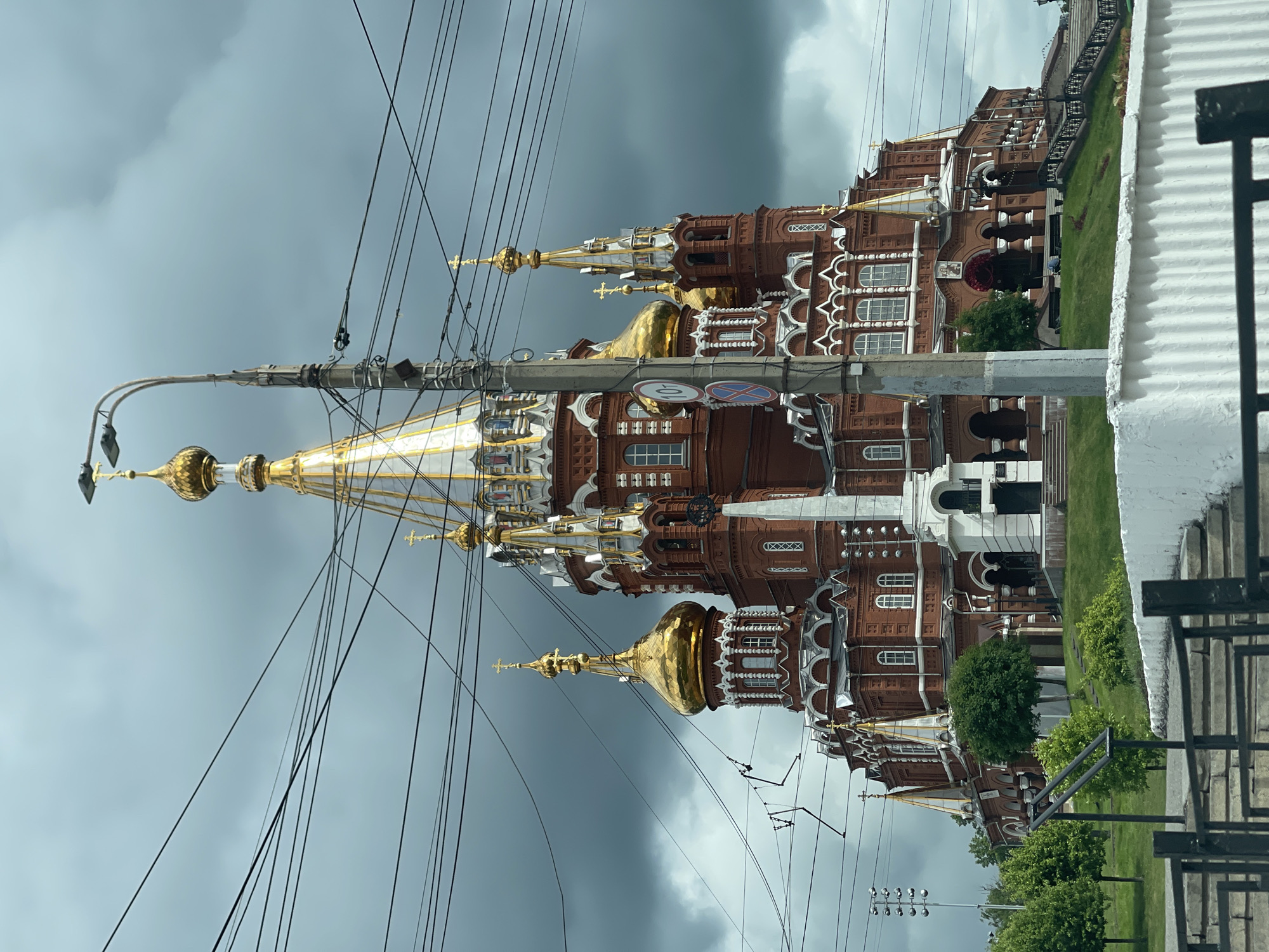 The image depicts St. Michael's Cathedral in Izhevsk, Russia, under a cloudy sky. The cathedral is characterized by its red brick facade, white trim, and prominent golden domes. A multitude of power lines intersect the foreground view of the cathedral and a light pole is present. The cathedral is situated next to a grassy area with trees and a partially visible white wall or building. A section of iron fence runs parallel to the grassy area. Several signs attached to the utility pole are visible near the top of the frame.