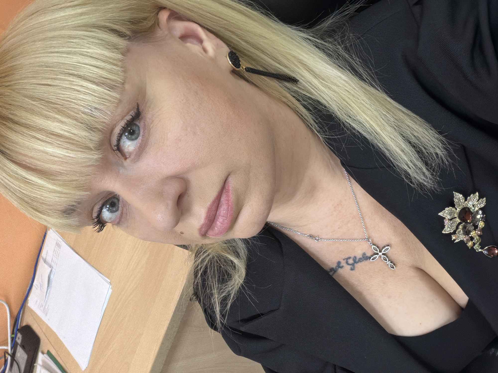 A medium, tilted, indoor close-up shot features a fair-skinned woman with blond, straight hair styled with blunt bangs, wearing black earrings and a black blazer. She is looking directly at the camera. A silver necklace with a cross pendant hangs around her neck above a tattoo on her chest. A decorative brooch is pinned to the blazer. A desk with papers and a phone is visible in the lower left corner. The location is unspecified, and cannot be identified as Chaikovsky, Russia from the provided image.