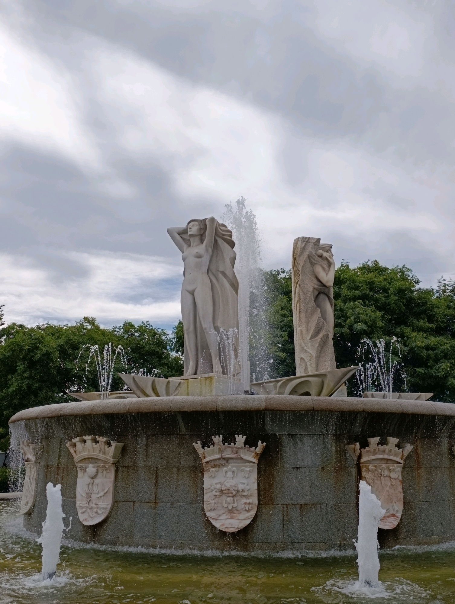 The image shows a large, ornate fountain with sculptures in Setúbal, Portugal. The fountain is made of stone and features two prominent nude female figures. The figure on the left is standing with her hands behind her head, draped in flowing fabric. The figure on the right is partially obscured by a textured stone slab and appears to be embracing herself. Water sprays from multiple points around the fountain, creating a dynamic and lively effect. The base of the fountain is decorated with several heraldic crests. Behind the fountain, lush green trees form a backdrop against a cloudy sky. The overall scene suggests a public park or plaza in the city.