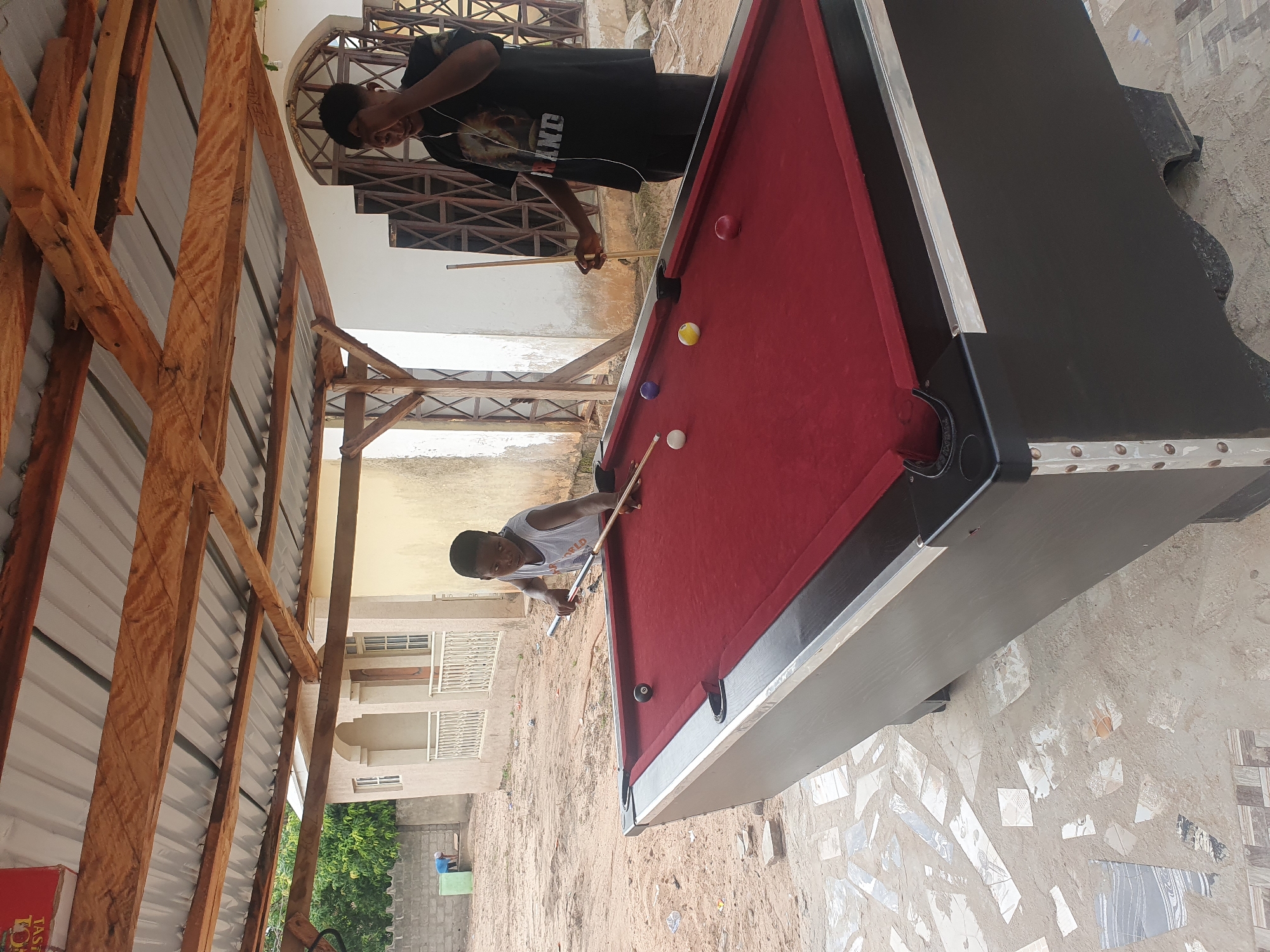 In Lokoja, Nigeria, two young males are engaged in a game of pool under a covered outdoor area. The area is paved with a mosaic of broken tiles on the ground, and a sandy area is visible in the background. A dark-colored pool table with a red felt surface dominates the foreground. One boy, wearing a light gray tank top, stands to the left, poised to strike a billiard ball with a cue. The other boy, in a black t-shirt, stands to the right, holding a cue and appears to be observing the game. Several billiard balls are scattered on the table. In the background, a building with a white facade and decorative metalwork is visible, along with some trees and a brick wall. The overall scene suggests a casual recreational activity in an outdoor setting.