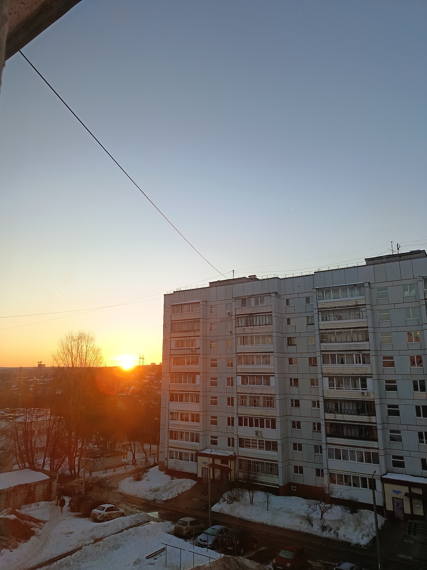 An outdoor, high-angle view captures a residential area in Chaikovsky, Russia, at sunset during winter. The clear sky, transitioning from light blue at the zenith to a bright orange-yellow at the horizon, dominates the upper portion of the frame. The setting sun, a prominent orange disc, is low on the left horizon, partially obscured by distant buildings and bare trees, casting long shadows and a strong golden hue across the scene. A faint white contrail is visible in the upper left sky, and a subtle crescent moon is present in the upper right.
A multi-story, light grey concrete apartment building, approximately nine to ten floors tall, occupies the right half of the image. Its facade features numerous rectangular windows, many with enclosed balconies, some reflecting the orange sunlight. The building's rooftop displays vents and antenna structures.
Below the main building, the ground level consists of paved surfaces, likely a parking lot or access road, largely covered in patches of dirty snow and slush. Multiple vehicles are parked: a dark sedan, a white sedan (possibly a taxi with yellow decals), a light-colored hatchback, a dark red car, and another dark sedan. A single individual, dressed in dark attire, walks on a snow-covered path near the parked cars. A small, single-story structure with a purple door and an illegible sign is integrated into the ground floor of the main apartment building.
To the left of the main building, a neighborhood extends into the distance, characterized by lower residential structures, bare deciduous trees, and substantial snow cover. Utility poles and power lines are visible on the distant horizon. A thick, dark cable runs diagonally across the sky from the upper left corner, passing above the main apartment building. Additional thinner, horizontal utility lines are present above the apartment building's roofline. The immediate foreground shows a concrete edge, likely a window frame or balcony from the vantage point of the photographer.