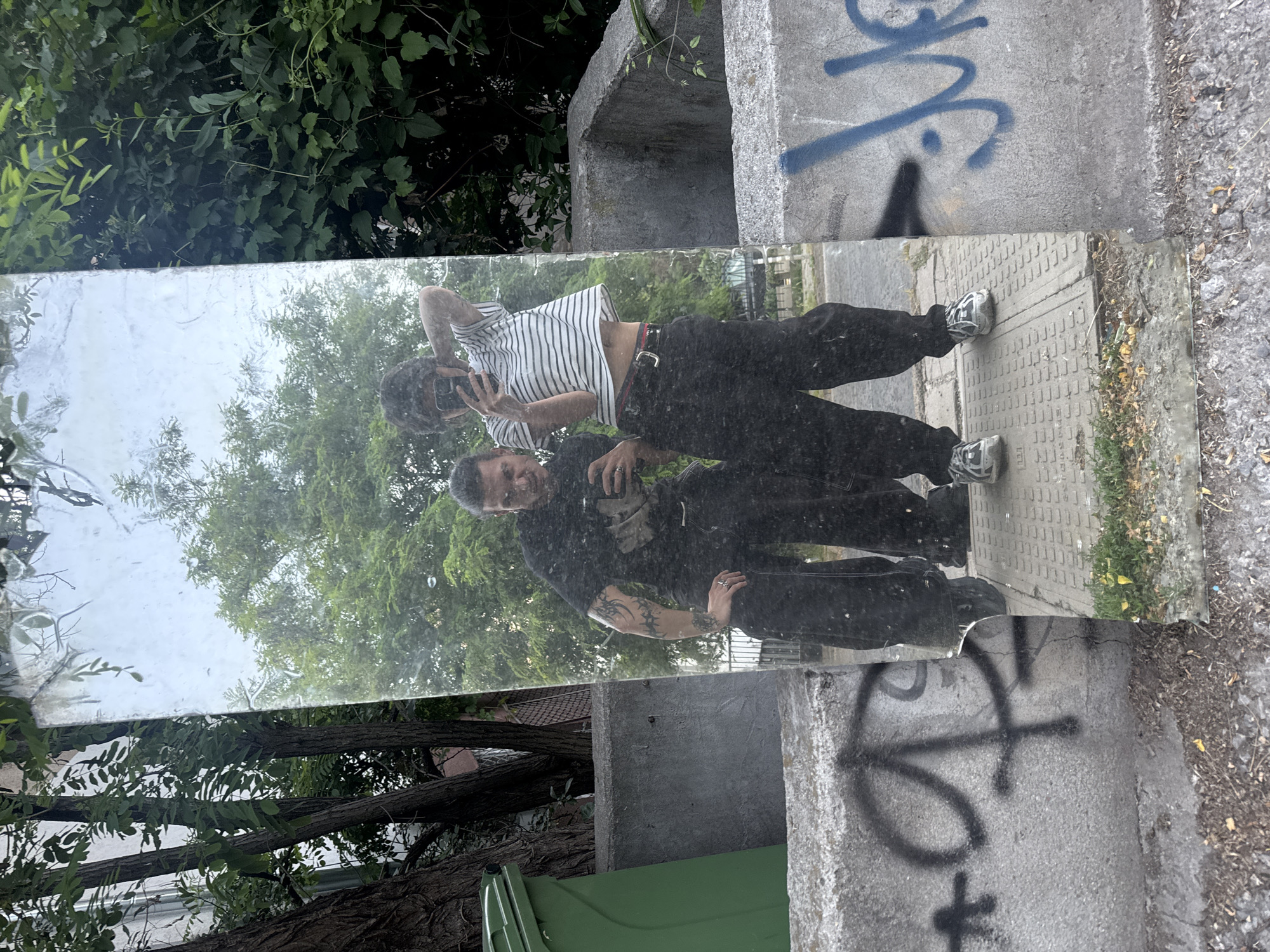 Two individuals are reflected in a tall, rectangular mirror placed vertically against a concrete barrier. The mirror is positioned outdoors, with trees and foliage visible in the background, suggesting an urban setting. The ground in front of the mirror is a mix of dirt, gravel, and a paved walkway with a textured surface.
The individual on the left is wearing a black t-shirt with a graphic print, black pants, and dark shoes. They are holding a smartphone in their right hand and looking at the camera. They have tattoos on their left arm. The individual on the right is wearing a striped t-shirt, black pants with a red-striped belt, and athletic shoes. They are also holding a smartphone and adjusting their hair with their left hand.
The concrete barrier in the foreground has graffiti on it, including black and blue markings. To the left of the mirror, a green garbage bin is partially visible. The overall scene appears to be an informal snapshot taken by the individuals themselves.
Based on visual cues such as the style of graffiti and the urban environment, it is not possible to definitively pinpoint the location within Sofia, Bulgaria.