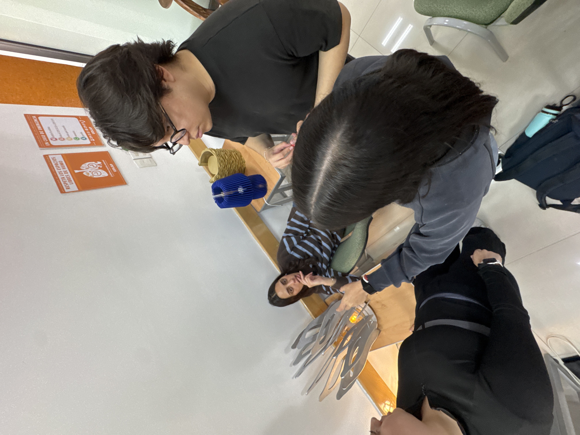 This image, taken in San Andrés Cholula, Mexico, depicts four individuals gathered around a light-colored wooden table in an indoor setting. The environment appears to be a university building, likely a common area, workshop, or exhibit space, confirmed by the "UDLAP" logo visible on a wall sign.
Two people, a male with dark hair and glasses (top left) and a female with dark hair (top right), are leaning over the table, intently focused on objects displayed. A third female (middle right), with long dark hair and wearing a blue and brown striped shirt, is seated at the table, looking towards the camera with a contemplative expression, her finger near her mouth. The fourth person (bottom), dressed in dark clothing, is also engaged with the table, with only part of their head and upper body visible.
On the table are several distinct objects: a light brown, cylindrical item made of woven or coiled material, a blue accordion-folded decorative piece, and an intricate metal sculpture composed of multiple curved, interlocking gray segments, which contains a small light source.
The background features white walls with an orange accent strip. Two framed signs are visible on the orange wall. The top sign is titled "EN CASO DE EMERGENCIA" (In case of emergency) and displays several circular icons indicating emergency procedures. The bottom sign reads "RESPETA LOS ESPACIOS LIBRES DE HUMO" (Respect smoke-free spaces) and features a no-smoking symbol over an image of lungs, along with the "UDLAP" logo. Green office chairs and a black backpack with a light blue water bottle are also partially visible in the background. The overall scene suggests a collaborative activity, possibly a design review, an exhibit viewing, or a workshop session. The lighting indicates daytime.