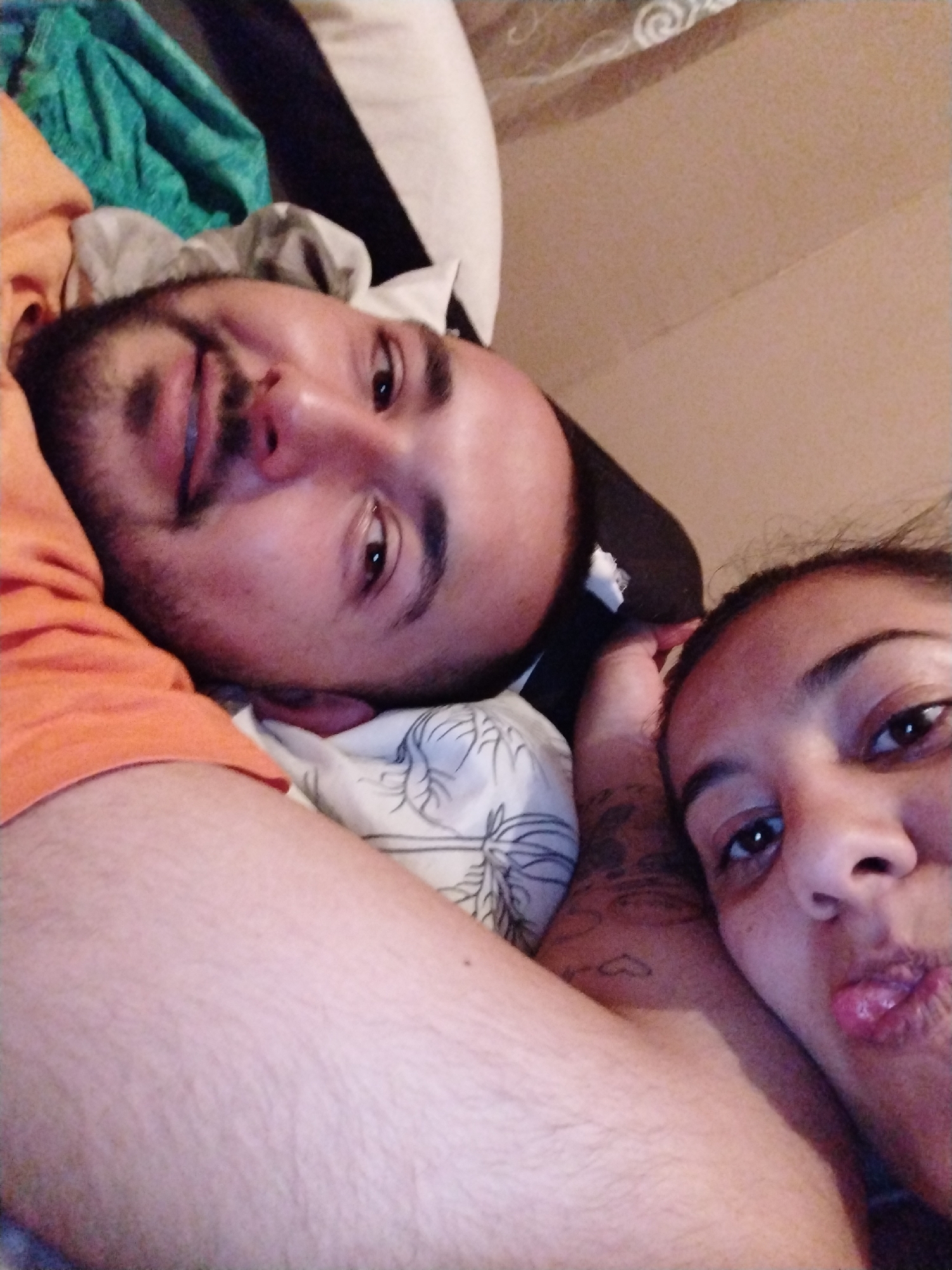 This is a selfie taken by a woman, likely indoors, of herself and a man lying down. The man is positioned to the left of the frame and slightly above the woman, with his head resting on a pillow. He has a beard and is wearing an orange shirt. The woman is on the right side of the frame, closer to the camera, and has dark hair. Her lips are slightly puckered. They appear to be lying on a bed with white bedding. A green cloth is visible in the upper-left corner of the frame. The location of the image is unknown.