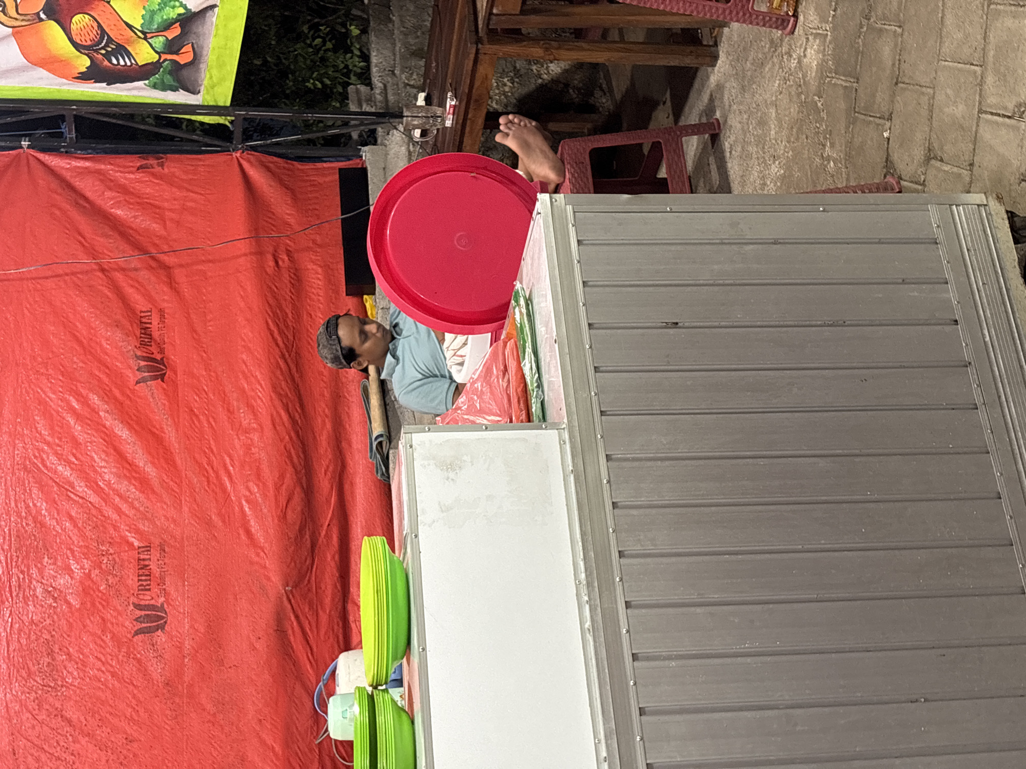In Kuta Selatan, Indonesia, a young man wearing a grey baseball cap and a light blue polo shirt sits with his eyes closed, appearing to be resting. He is positioned behind a counter or food stall, in front of a large red tarp that serves as a backdrop. On the counter, several bright green plastic bowls are stacked. To the right of the man, a large, bright pink plastic lid is prominently displayed. His right leg is visible, resting on a red plastic stool, with his bare foot exposed. The scene takes place outdoors, on a paved area, with a wooden table and chairs visible in the background. A caricature of a bird is depicted on a banner to the right. The lighting suggests it is evening.